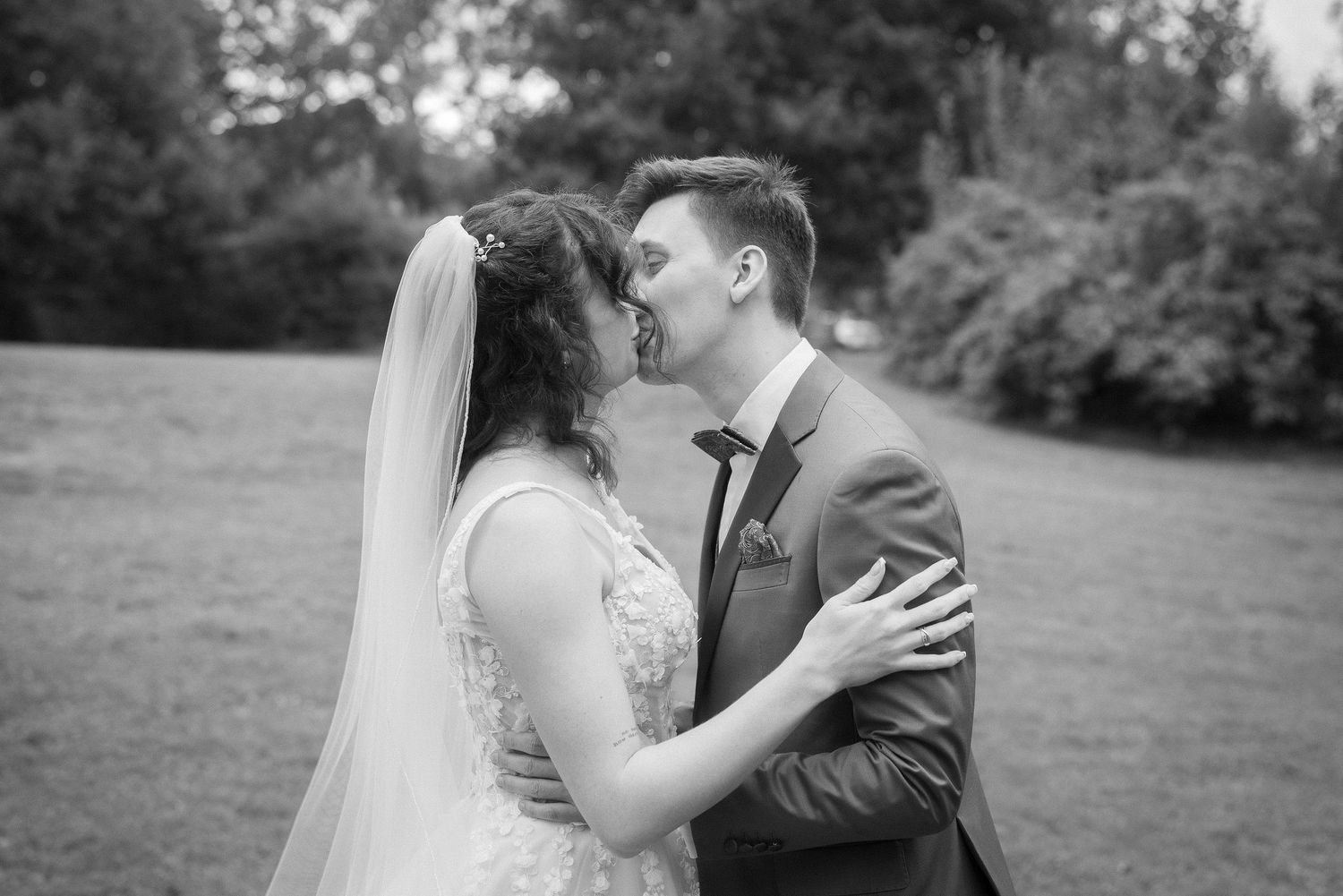 Black and white photo of a bride in a lace gown and veil kissing her groom in a suit on a grassy lawn surrounded by trees.