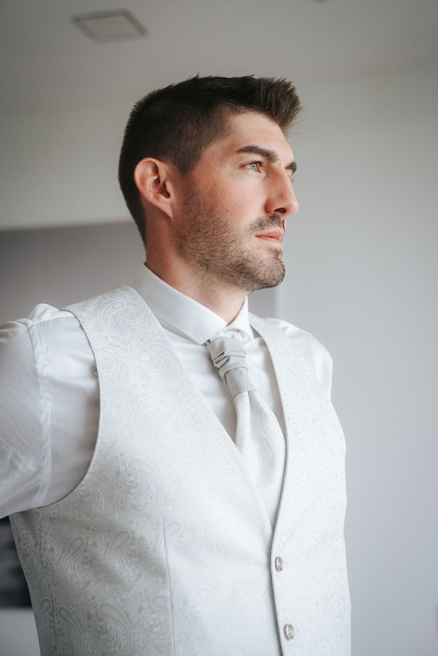 Groom in a white paisley vest, white shirt and silver ruche cravat gazing out a window while getting ready for his wedding.