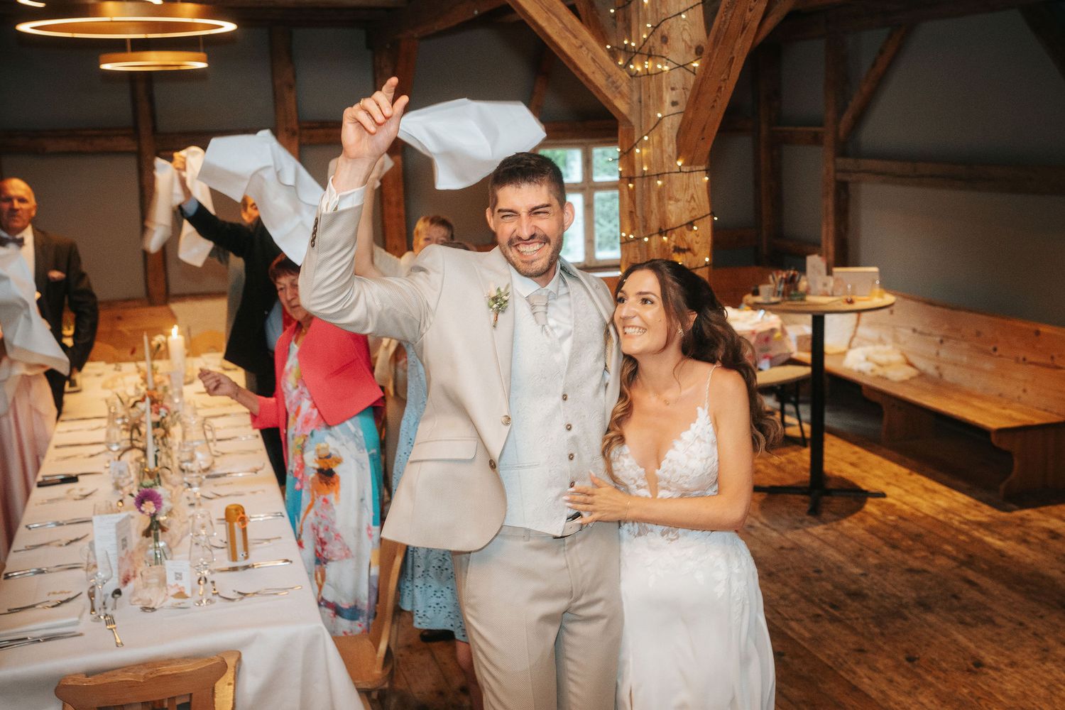 Smiling groom in a beige suit waves a napkin beside his bride in a lace gown during a rustic wedding reception with guests and set tables.