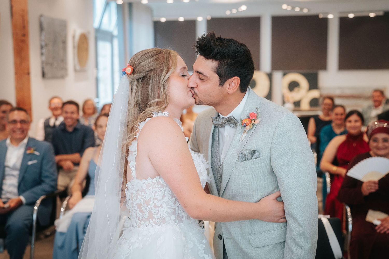Bride in a lace gown and veil kisses groom in a light gray suit during a wedding ceremony as seated guests look on.
