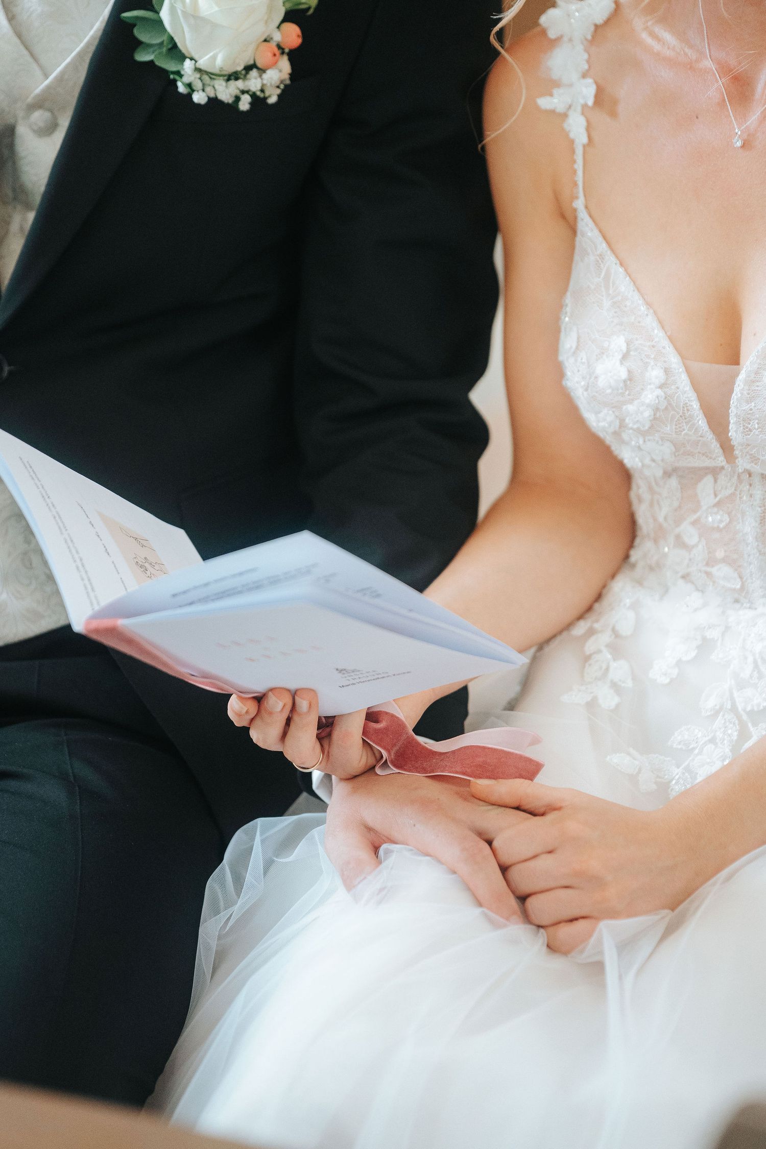 Bride in a white lace wedding dress and groom in a dark suit with floral boutonniere holding hands while reading a ceremony booklet.