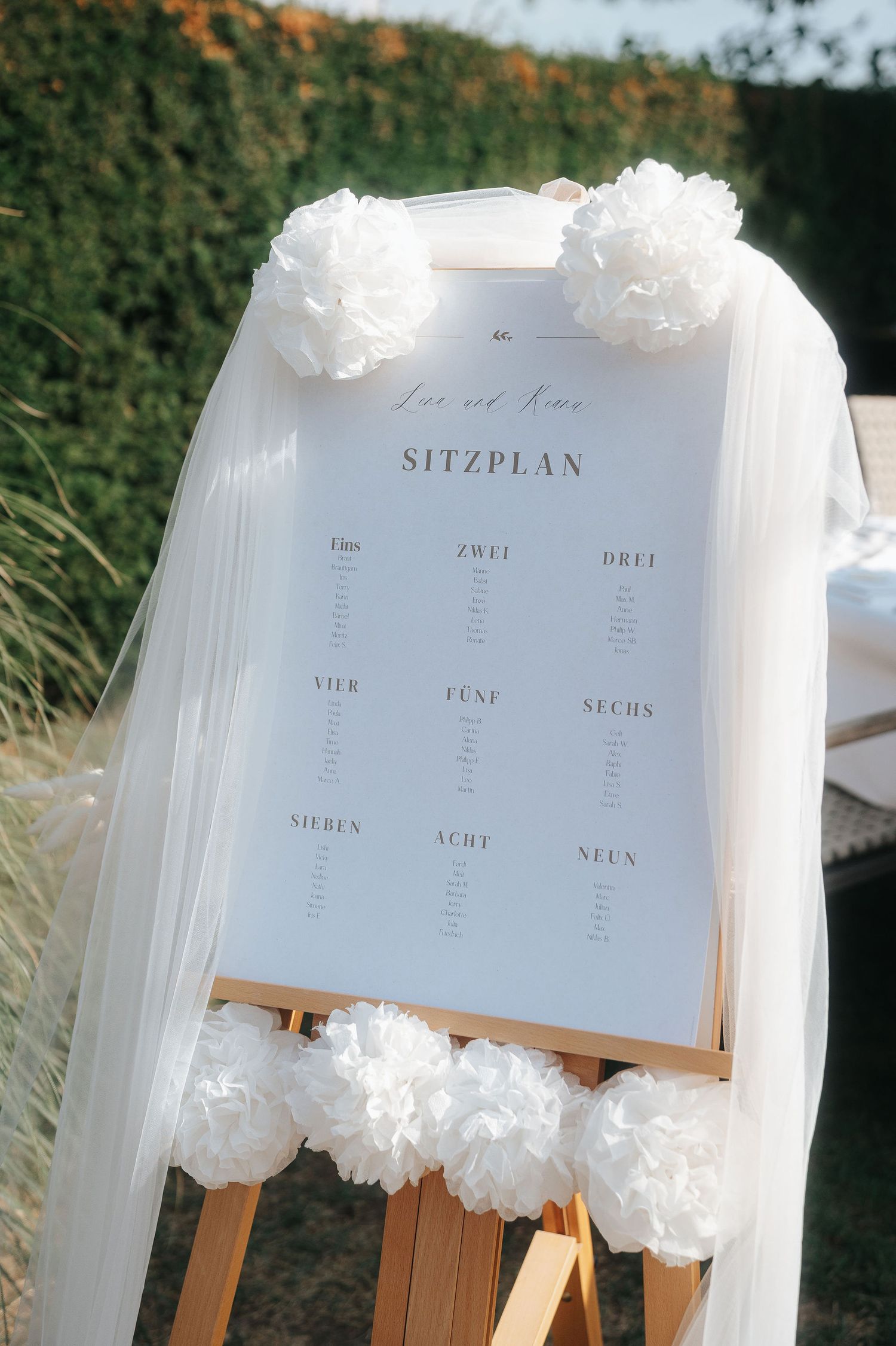 Wedding seating chart labeled Sitzplan on a wooden easel, decorated with white paper pom-poms and sheer tulle outdoors.