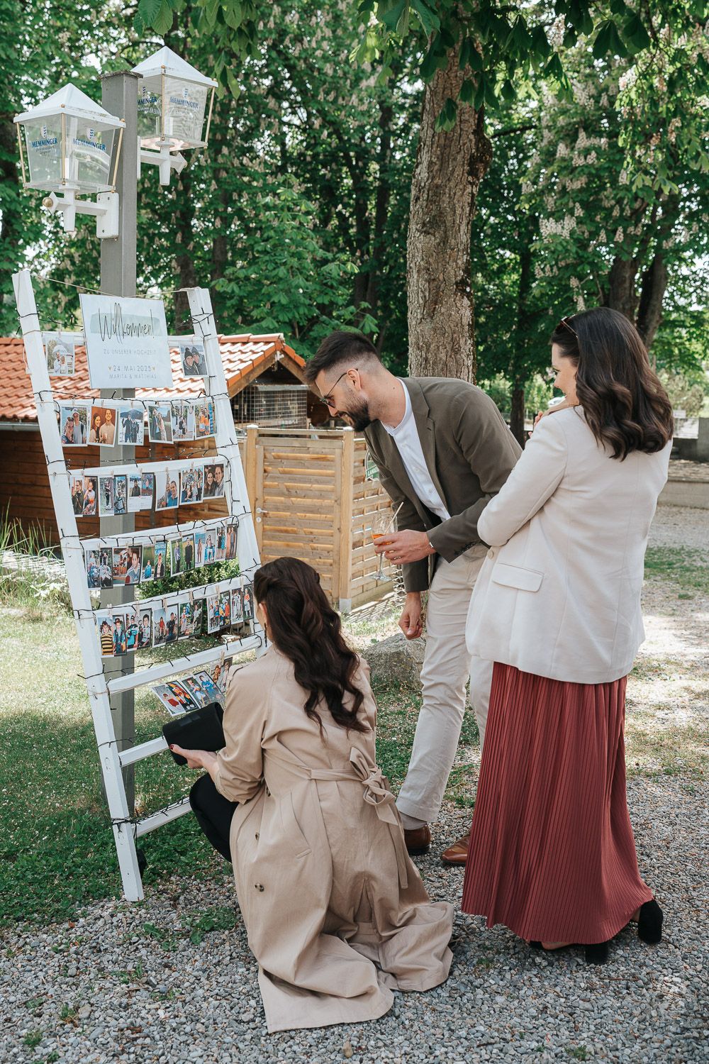 Group browsing photo display on white ladder shelf with decorative lanterns in outdoor garden setting.