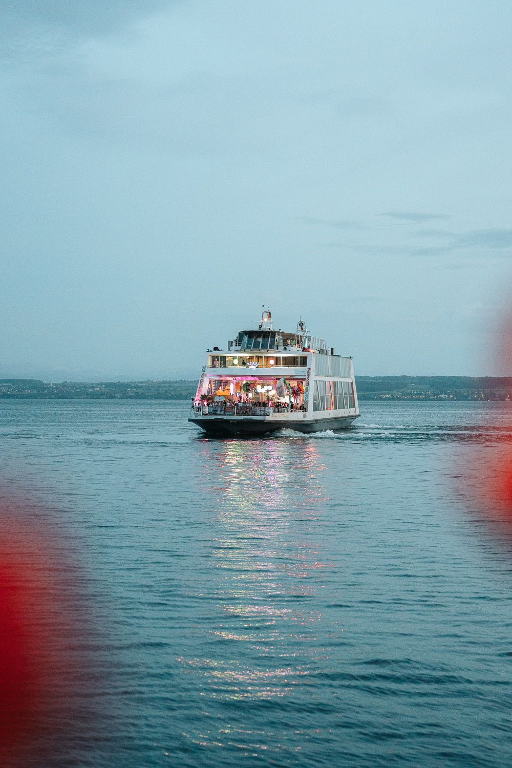 A passenger ferry boat cruises across calm blue waters with red tinted lens flare in the foreground.