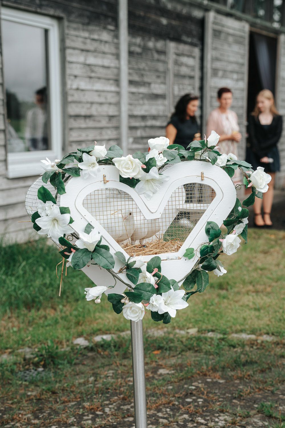 A heart-shaped wedding decoration stand adorned with white flowers and greenery stands in front of a rustic building.