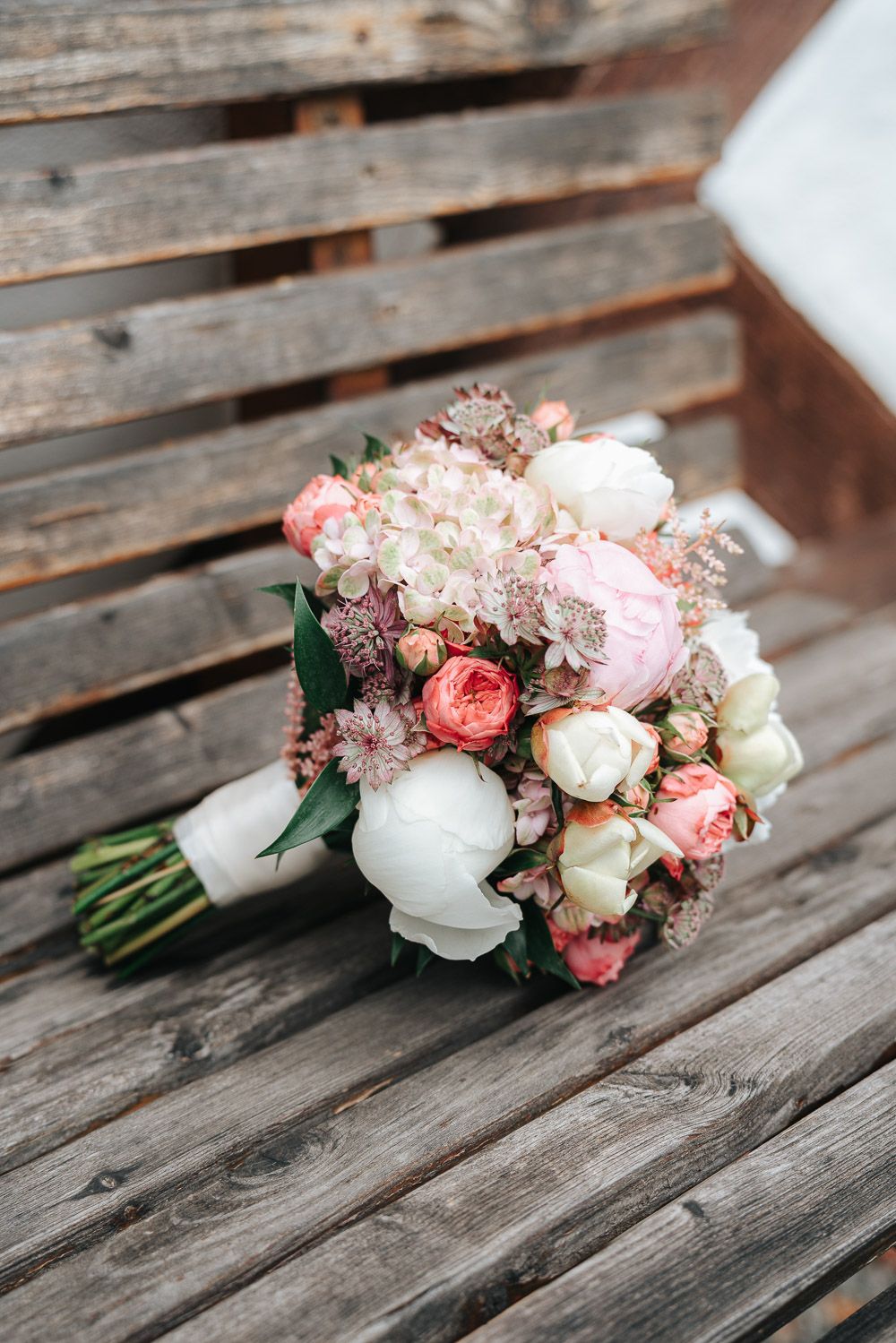A romantic bridal bouquet with white peonies, pink garden roses, and delicate blush flowers rests on rustic wooden planks.