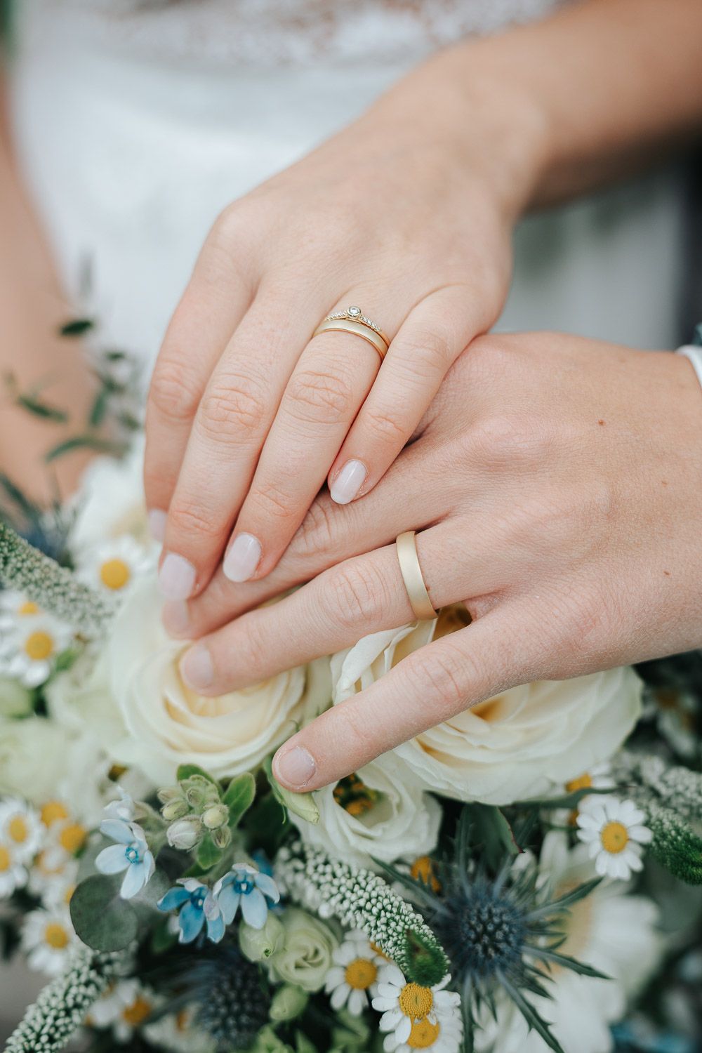 Close-up of hands with gold wedding bands resting on white roses and daisies in a soft floral arrangement.