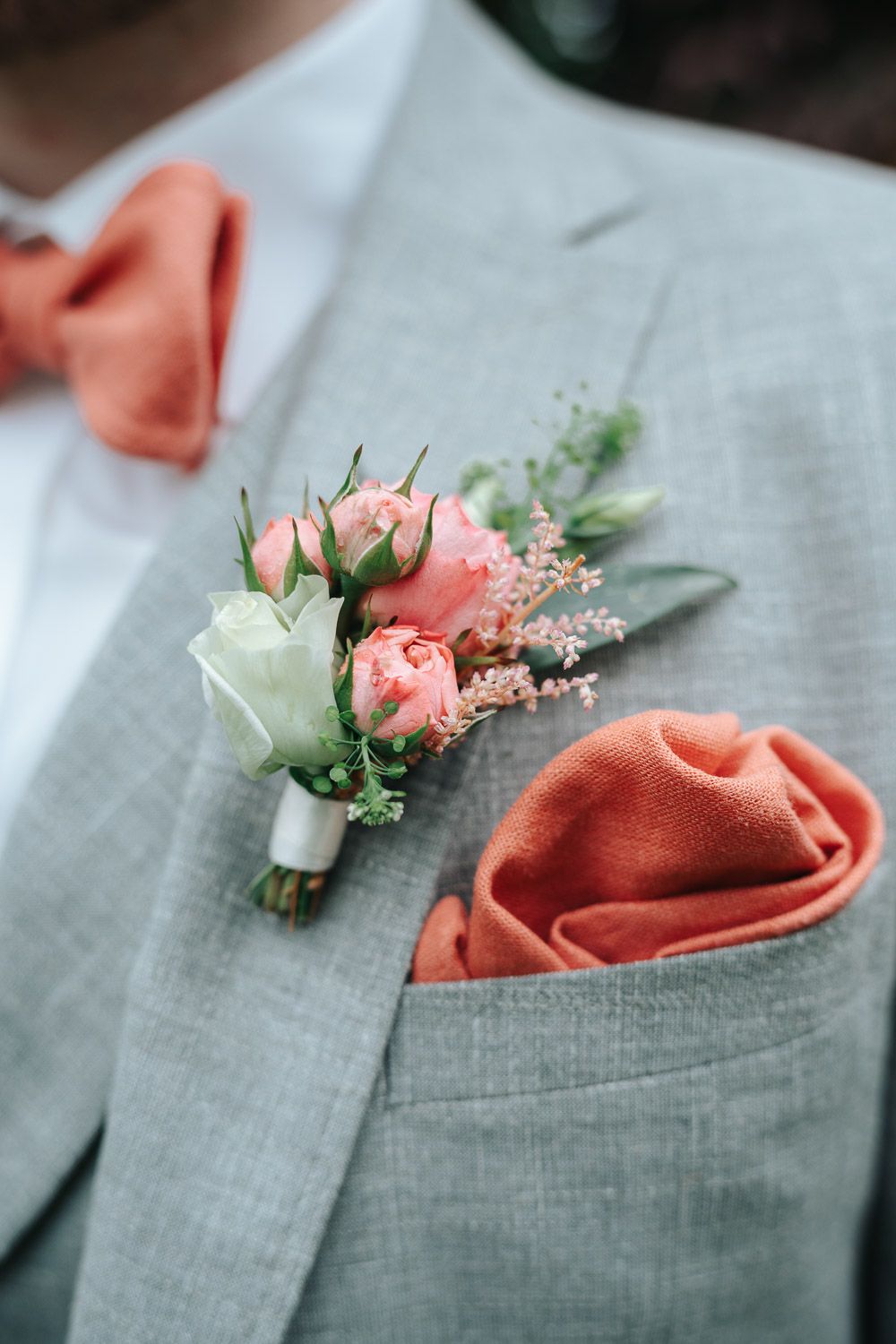 A coral rose and white floral boutonniere pinned to a light gray suit jacket with matching coral pocket square.