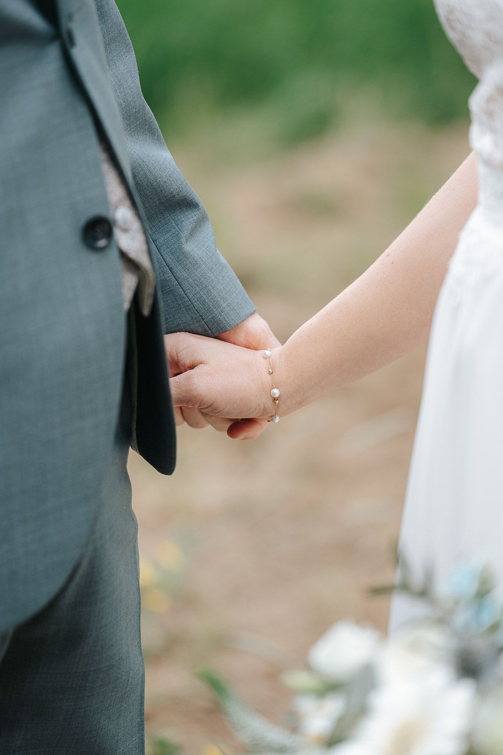 A couple holding hands wearing wedding attire - grey suit jacket and white dress against a soft green background.