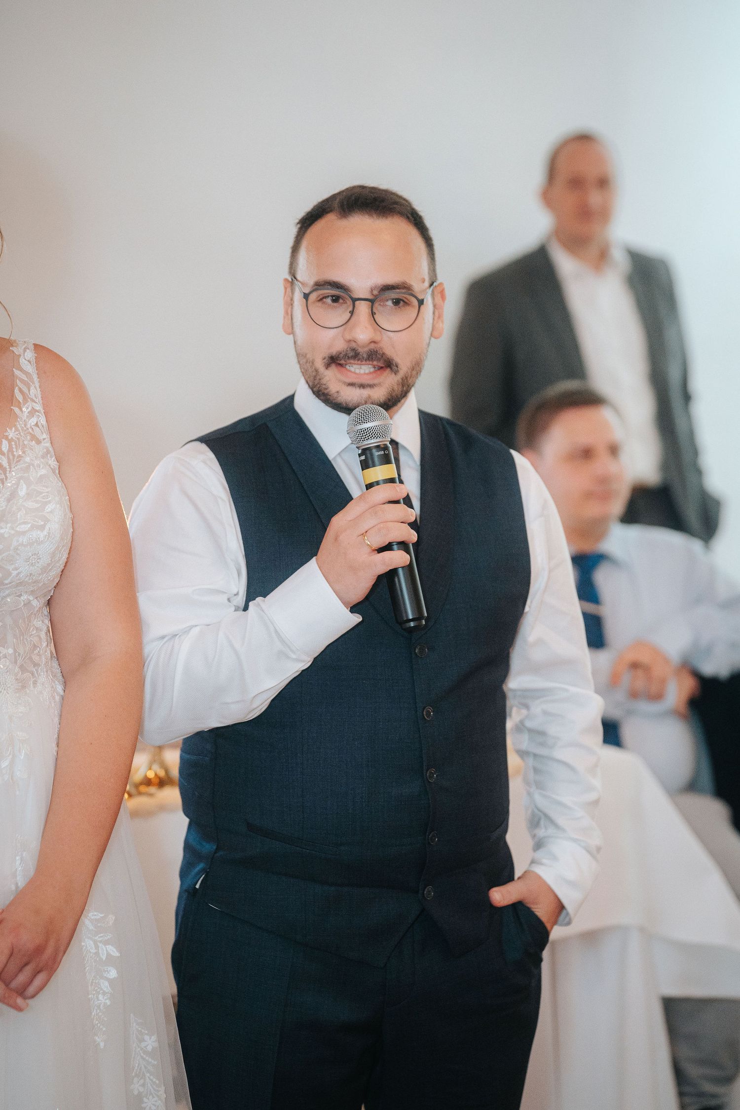 Man in glasses, navy vest and white shirt speaks into a microphone at a wedding reception while guests and a bride in lace look on.