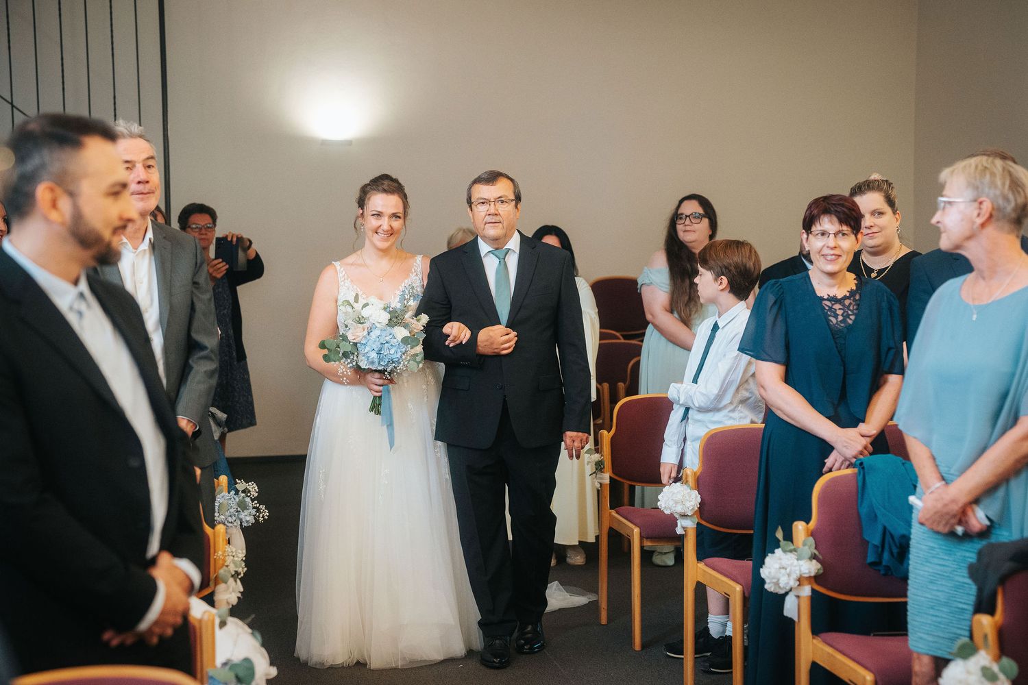 A bride in a white gown carrying a bouquet walks down the aisle with a man in a dark suit as wedding guests watch from seated rows.