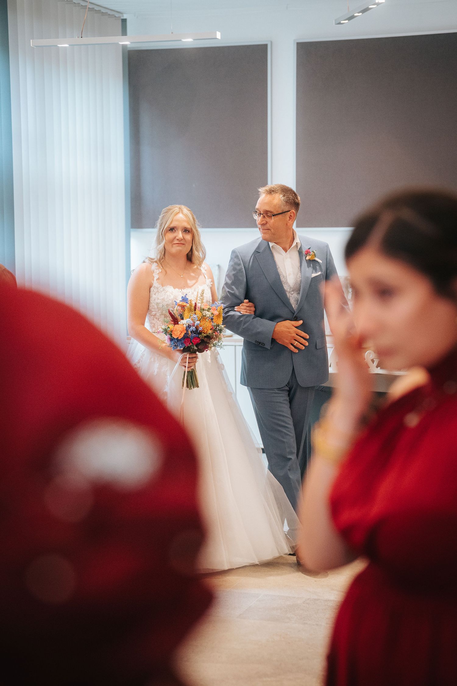 A bride in a lace wedding gown holding a colorful bouquet walks arm in arm with a man in a gray suit while a guest in red looks on.