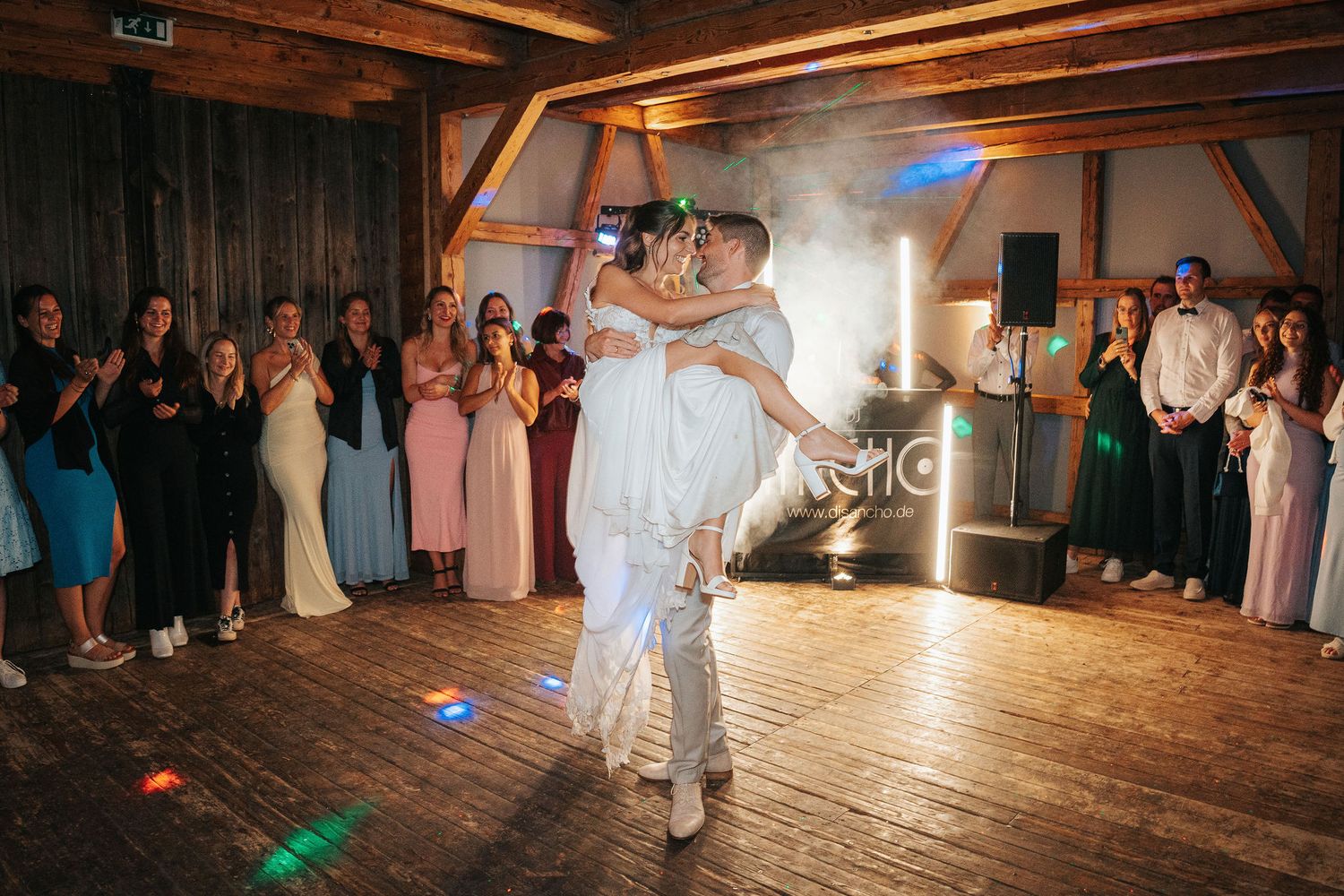 Bride and groom share their first dance on a wooden floor in a rustic barn venue as wedding guests watch and smoke fills the air.