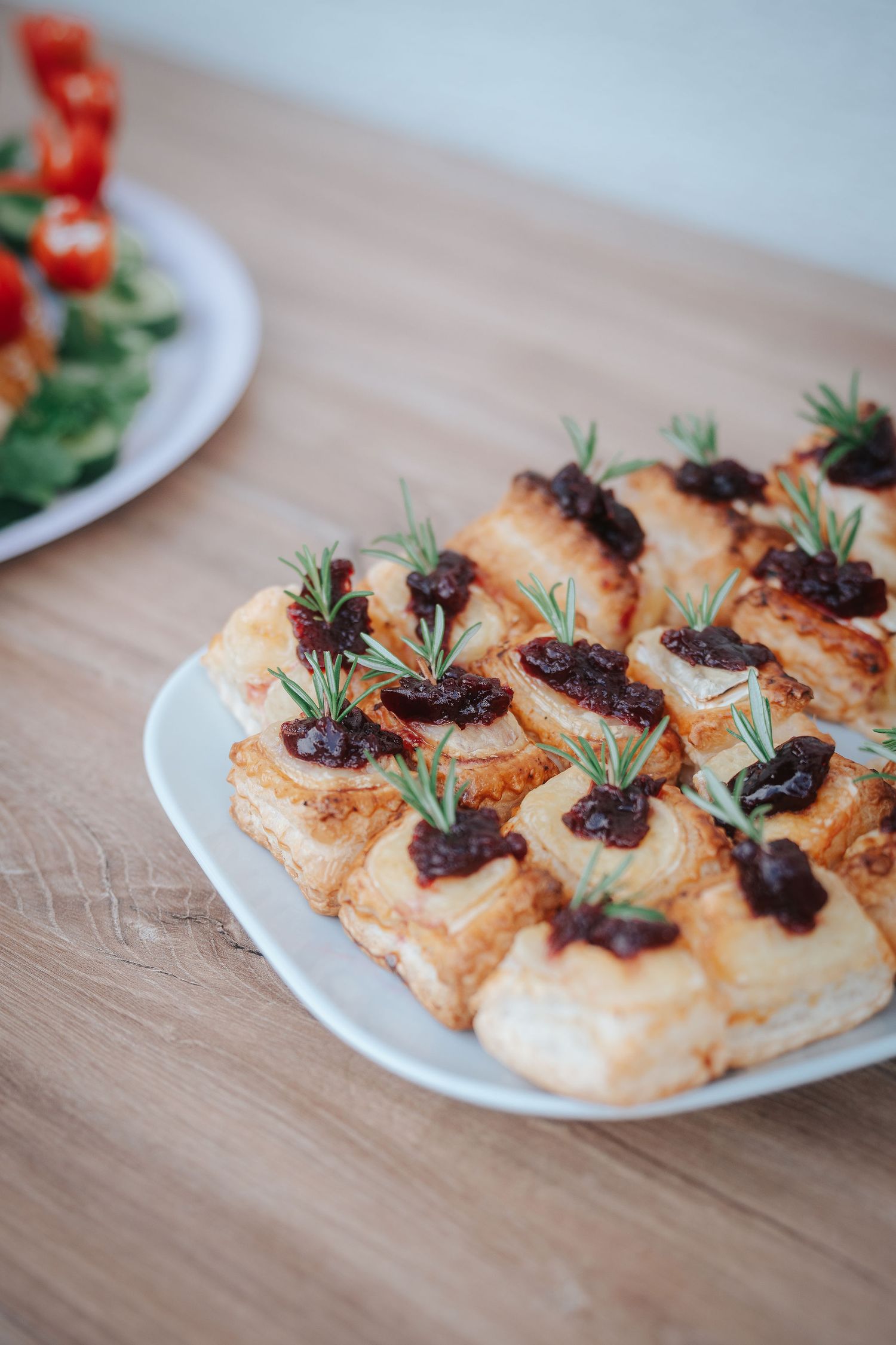 Puff pastry appetizers topped with dark berry jam and fresh rosemary sprigs on a white plate, served on a wooden table.