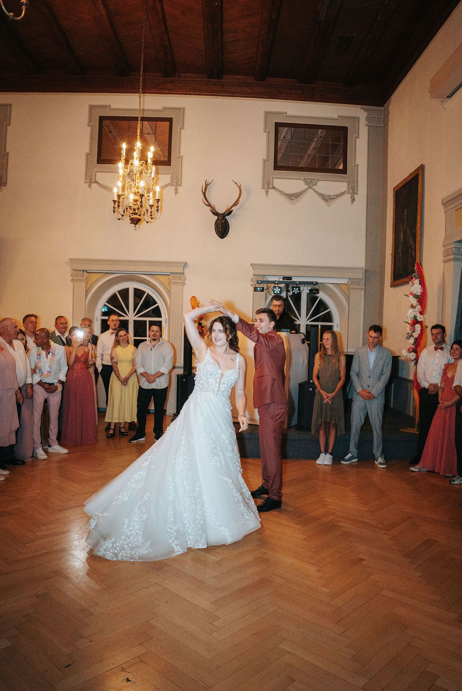 Bride in a lace gown and groom in a burgundy suit share a first dance on a wood floor as wedding guests watch in a historic hall.