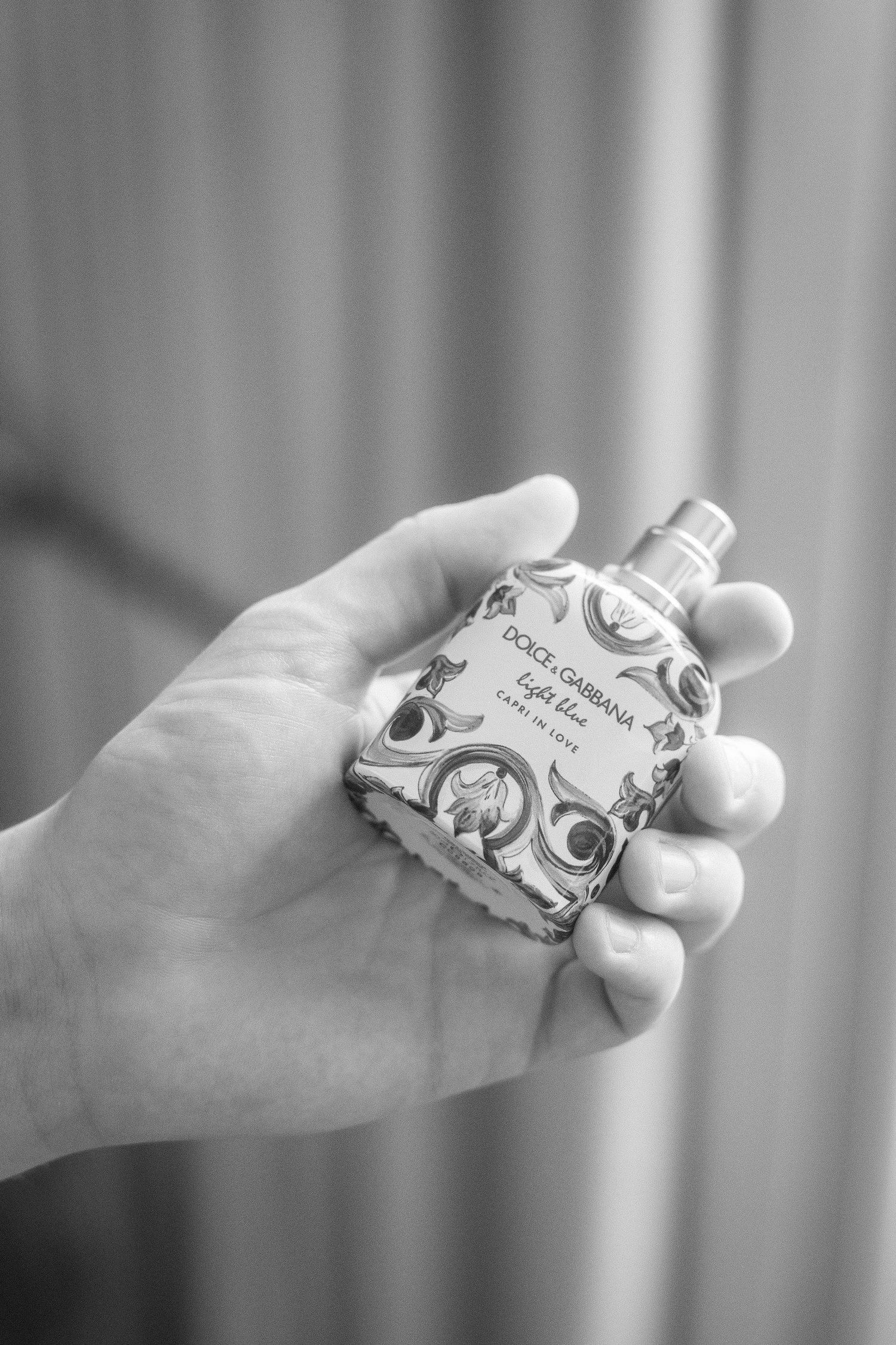 Black and white photo of a hand holding a Dolce & Gabbana Light Blue Eau de Toilette perfume bottle with ornate decorative patterns.