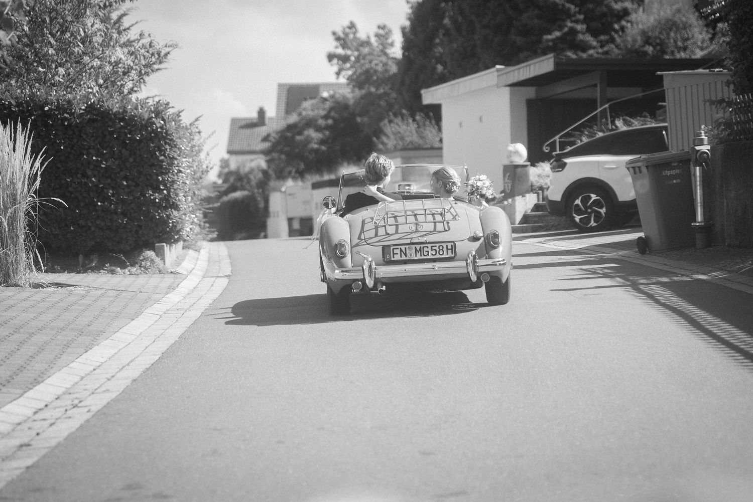 Black and white photo of a vintage convertible sports car with a license plate driving down a quiet residential street.