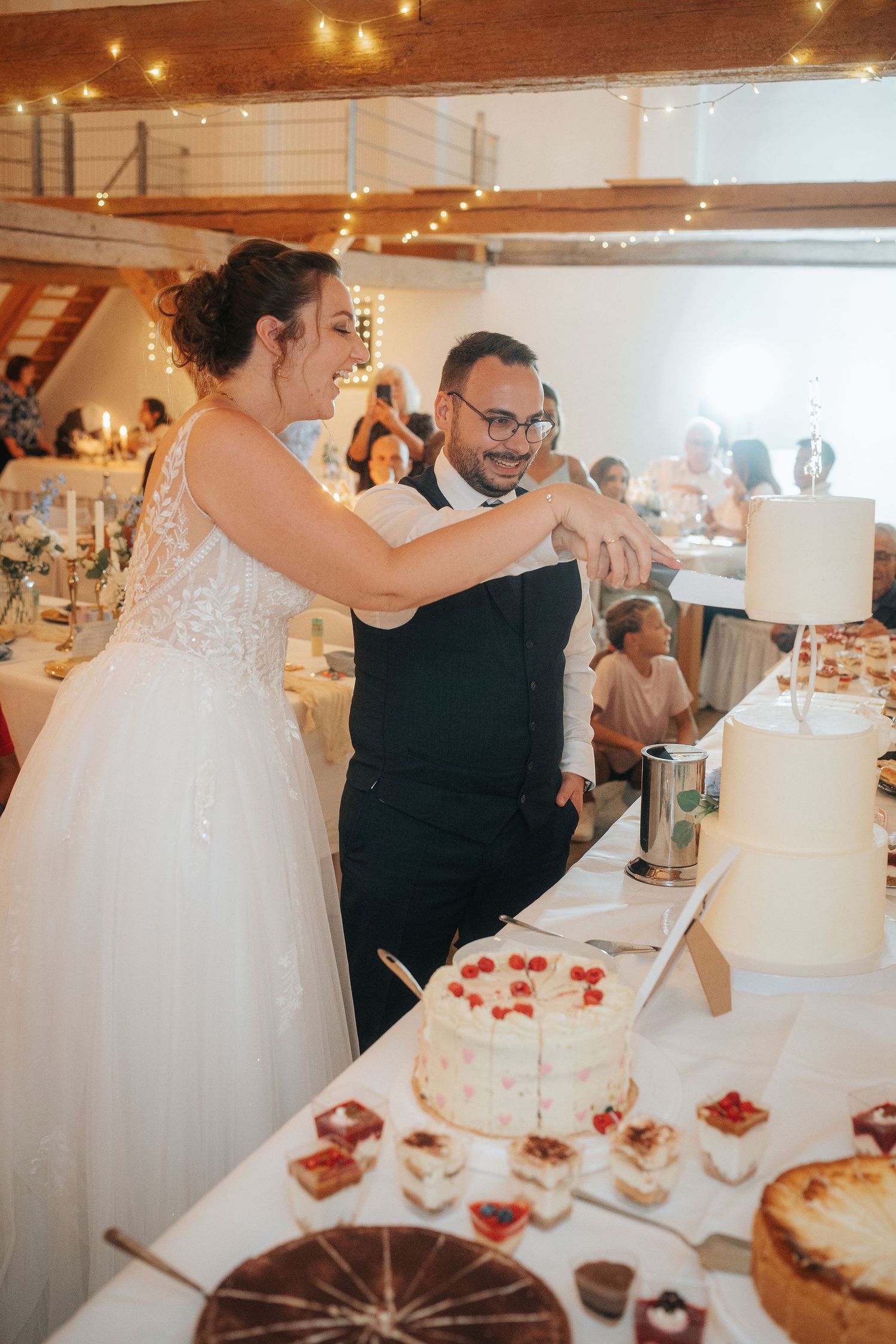 Bride and groom smiling as they cut their tiered wedding cake together at a rustic reception with a dessert table and guests nearby.