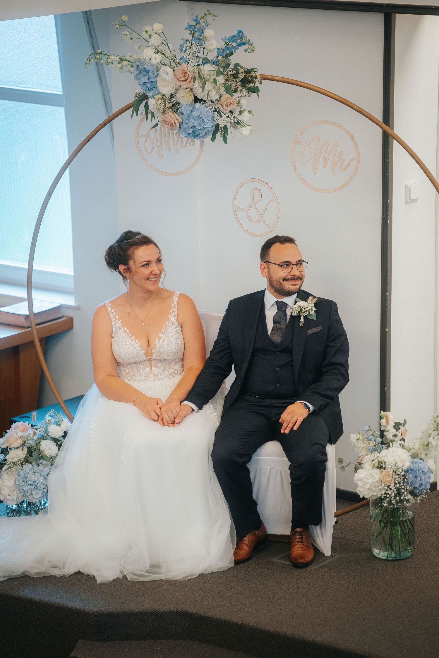 Smiling bride in a lace wedding gown and groom in a navy suit sit holding hands beneath a round floral arch with blue and blush flowers.