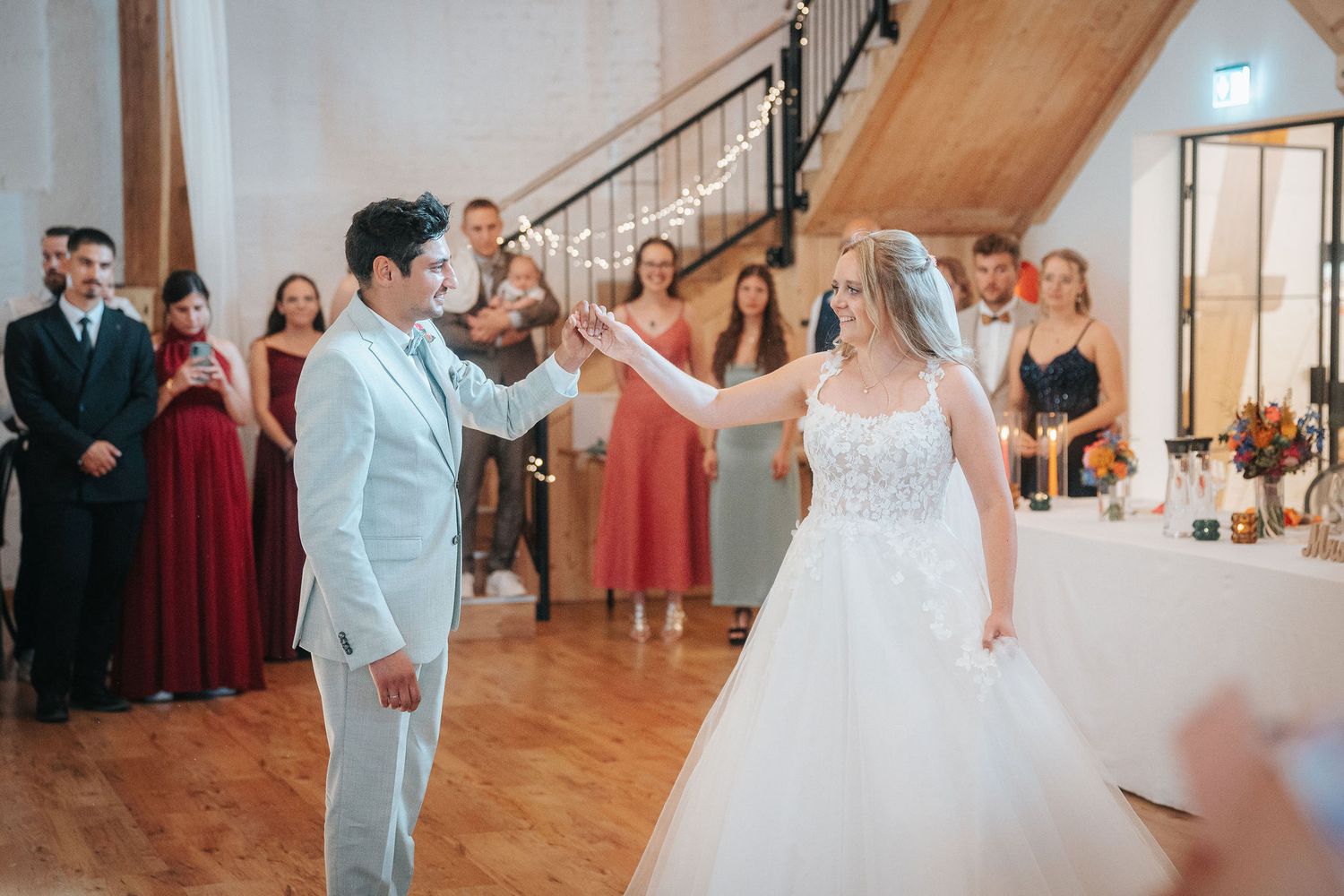Bride in a white lace gown and groom in a light blue suit share a first dance as wedding guests watch in a rustic hall.