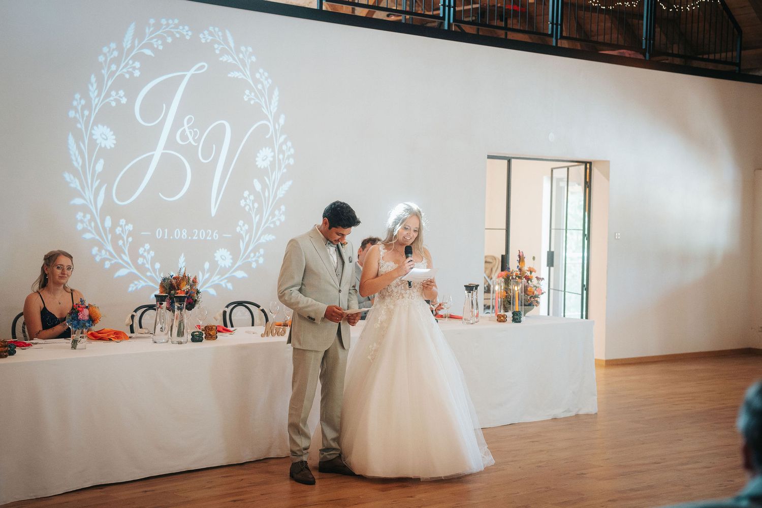 Bride in a white gown and groom in a gray suit give a wedding toast beside a decorated reception table under a monogram light projection.
