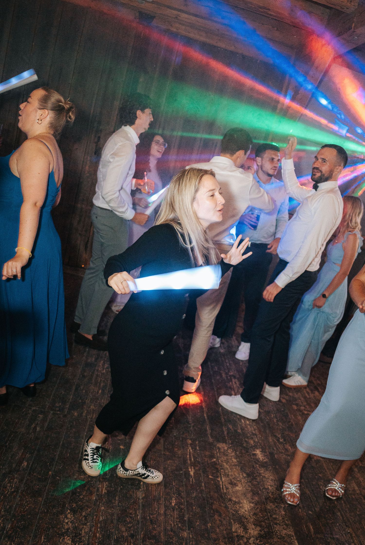 Wedding guests dance on a wooden floor under colorful party lights as a blonde woman in black dances with a glowing light stick.