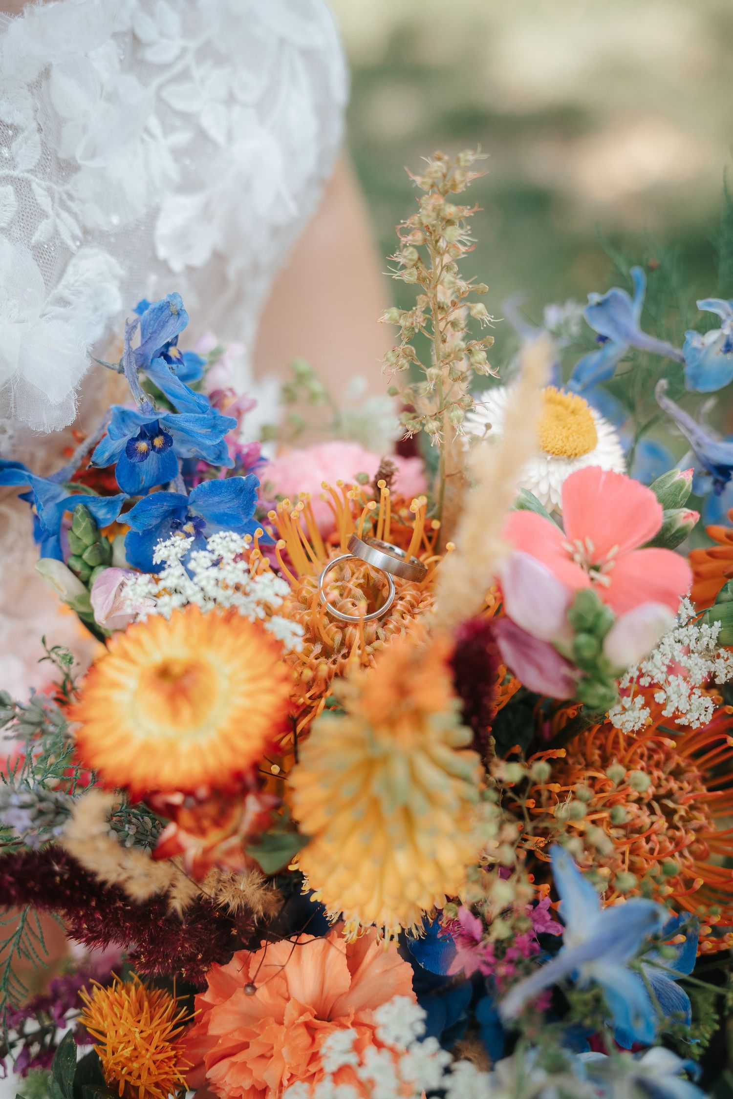 Bride in a lace gown holds a colorful wildflower bouquet of blue, orange and pink blooms with two wedding rings resting among the flowers.