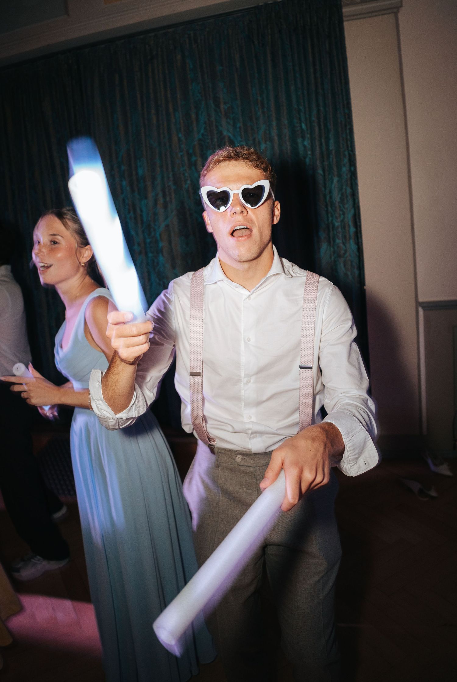 Man in heart-shaped sunglasses and suspenders dances with glow sticks at a wedding reception while a woman in a blue dress dances behind.
