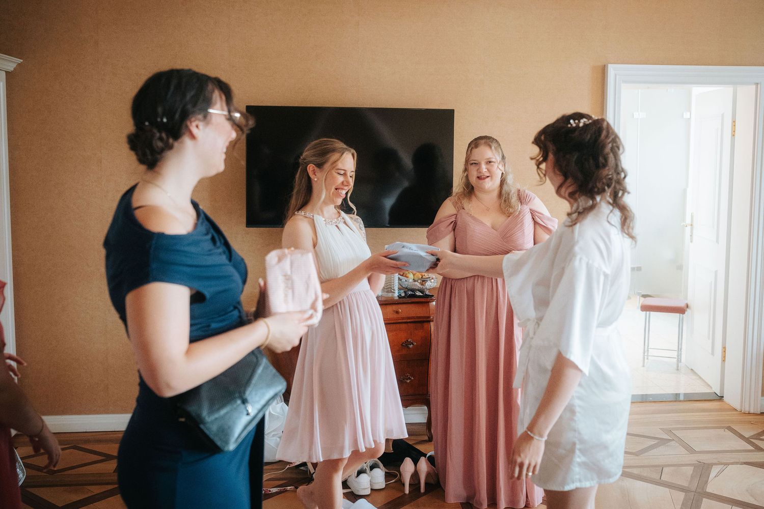 Four women in blush pink dresses and casual attire exchange gifts and smile together in a bright room while getting ready for a wedding.