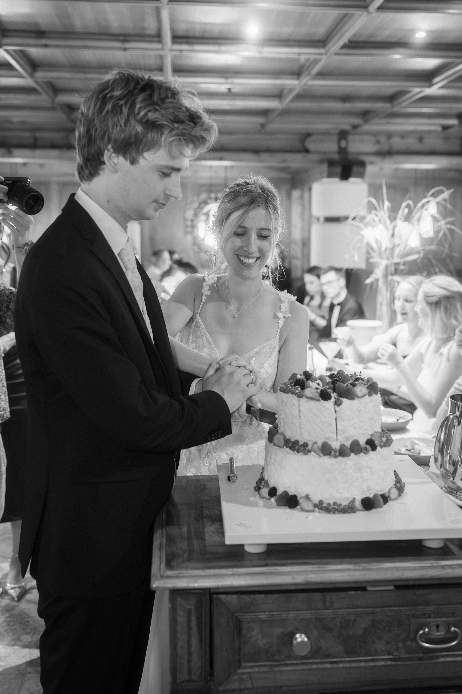 Black and white photo of a smiling bride and groom cutting their berry-topped wedding cake together at a indoor reception.