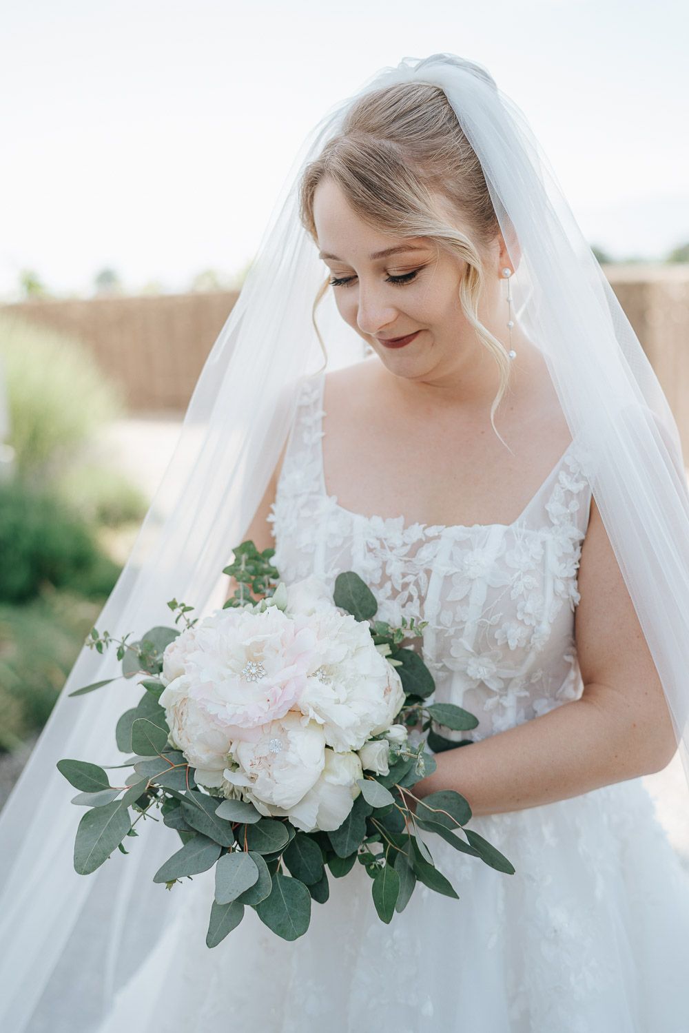 A bride in a white lace dress holds a romantic bouquet of pale pink flowers and eucalyptus.