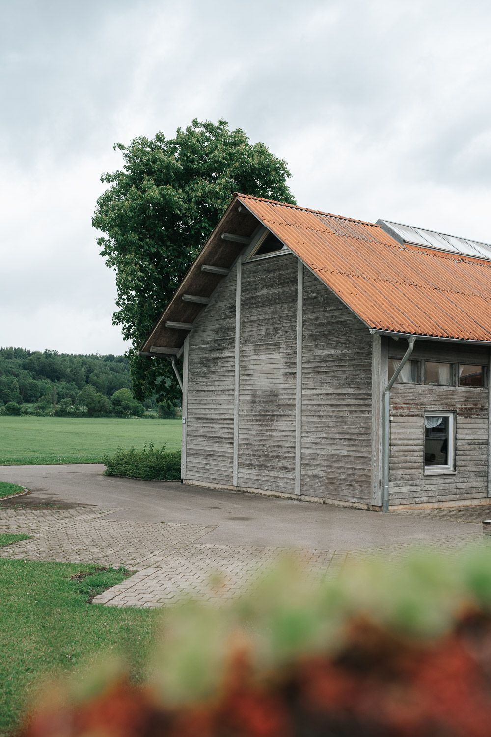 A series of rustic wooden barns with terracotta roofs stand against a cloudy sky in countryside setting.