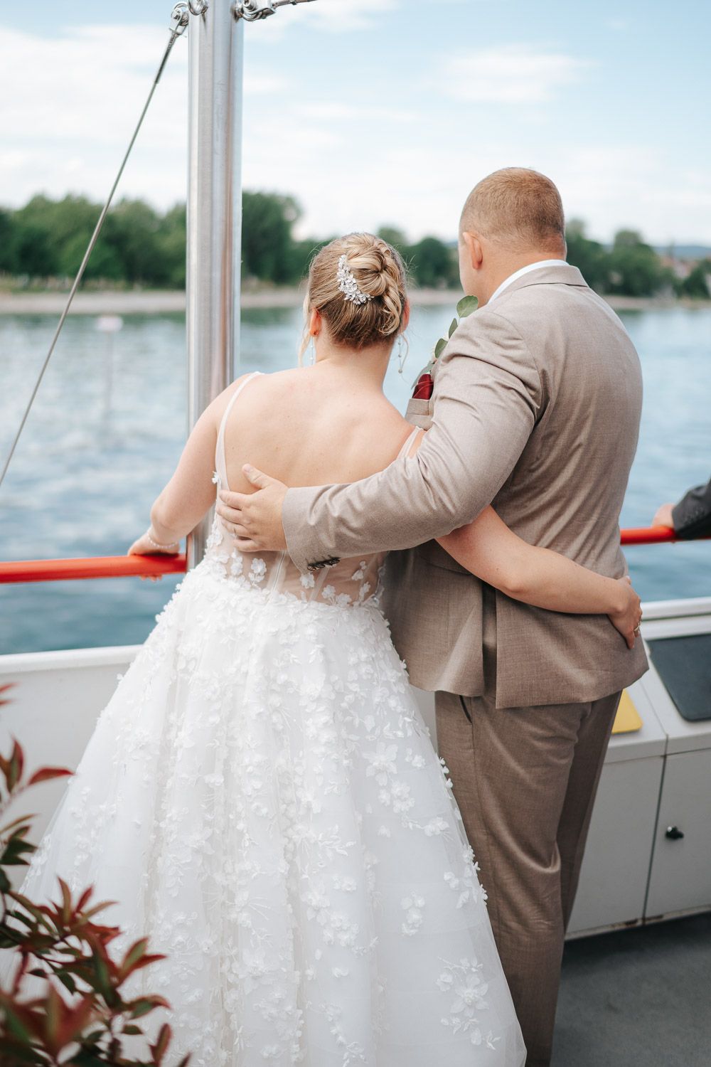 A romantic couple embraces on a boat deck during their wedding celebration, with bride in white gown and groom in tan suit.