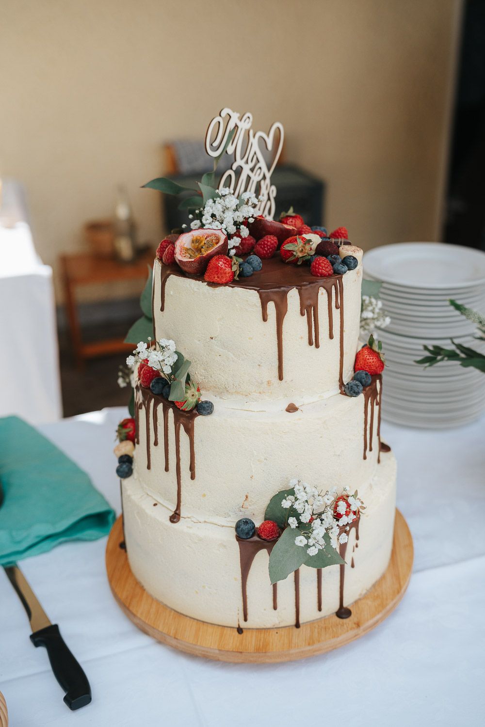 White frosted wedding cake with chocolate drip decorations and fresh berries displayed on wooden board from different angles.
