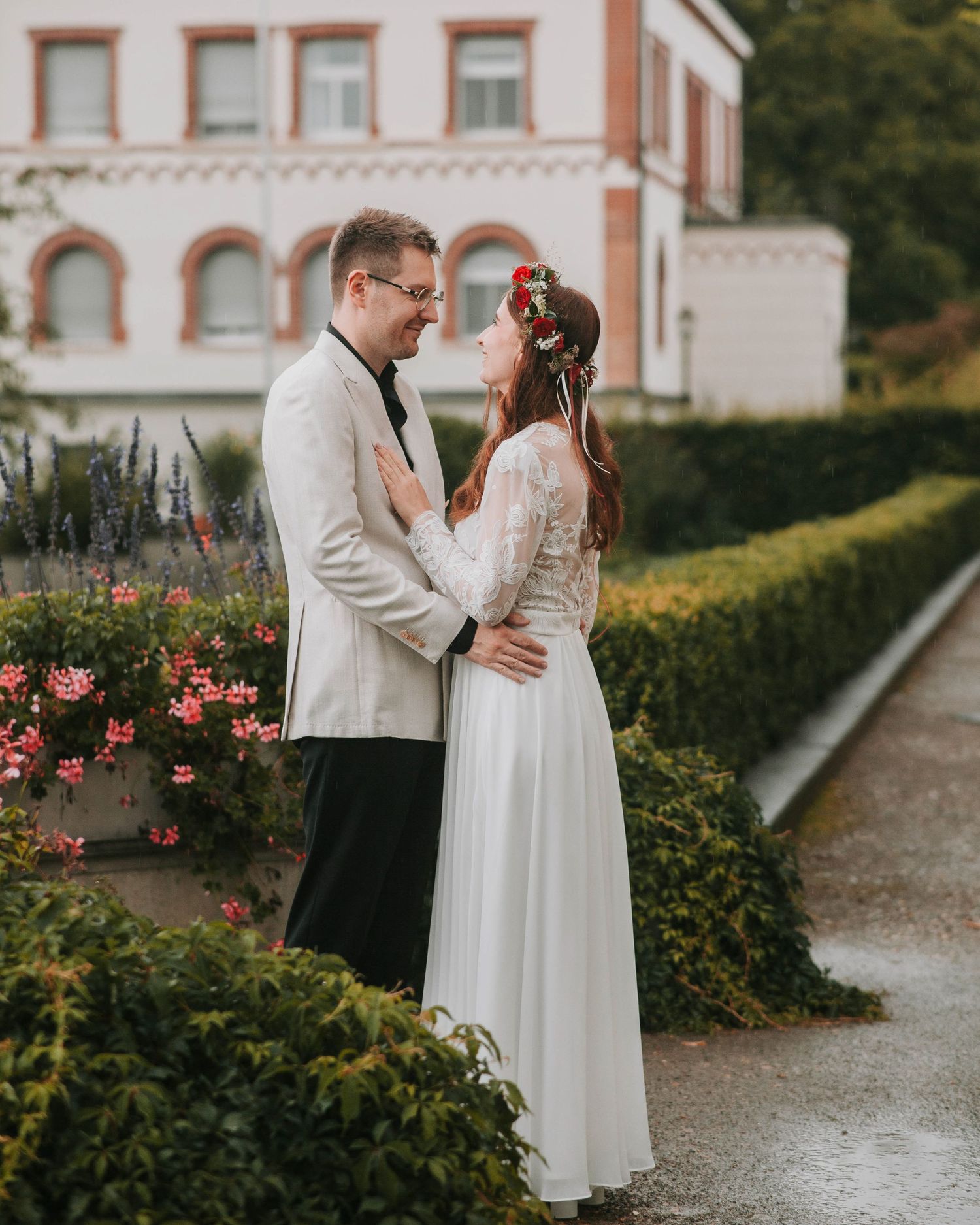 A couple in wedding attire shares intimate moments in a garden path lined with pink roses and white buildings.
