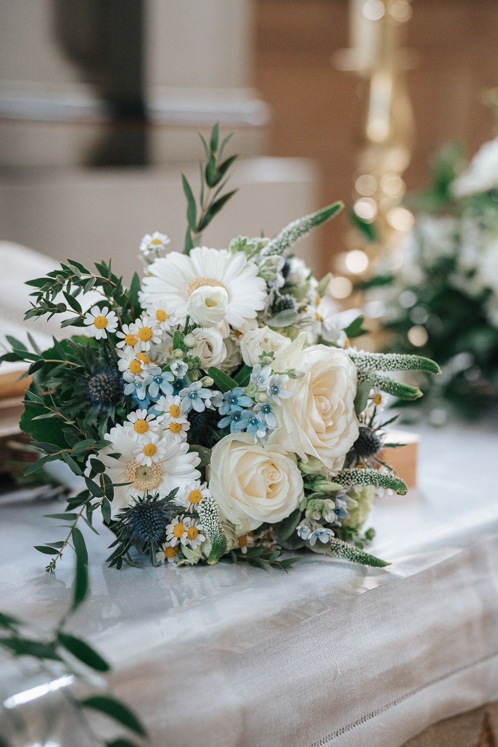 A romantic wedding bouquet with white roses, blue delphiniums, daisies and thistle arranged on a white tablecloth.