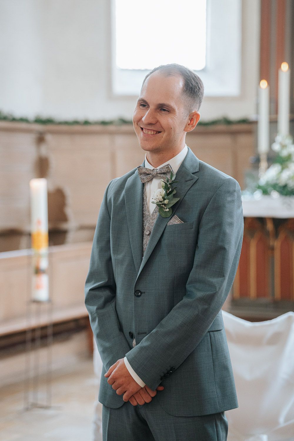 Groom in sage green suit with floral boutonniere smiles during wedding ceremony in sunlit church.