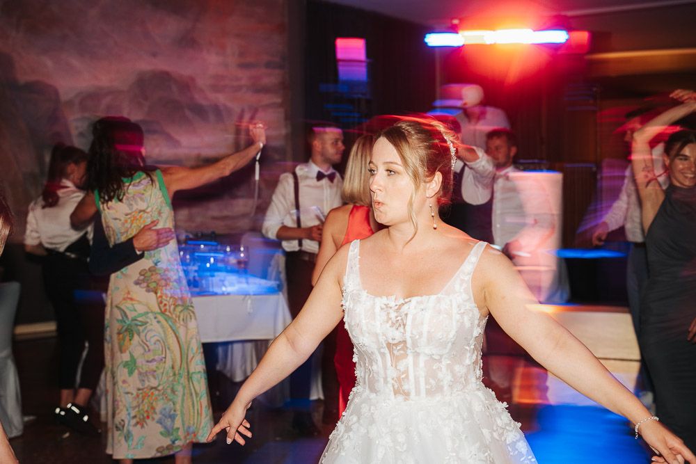 A person in a white bridal dress dances energetically on a nightclub dance floor under colorful lighting.