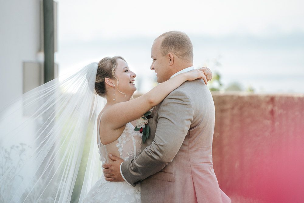 A joyful couple embrace lovingly during their wedding ceremony with a flowing white veil and soft beige suit.