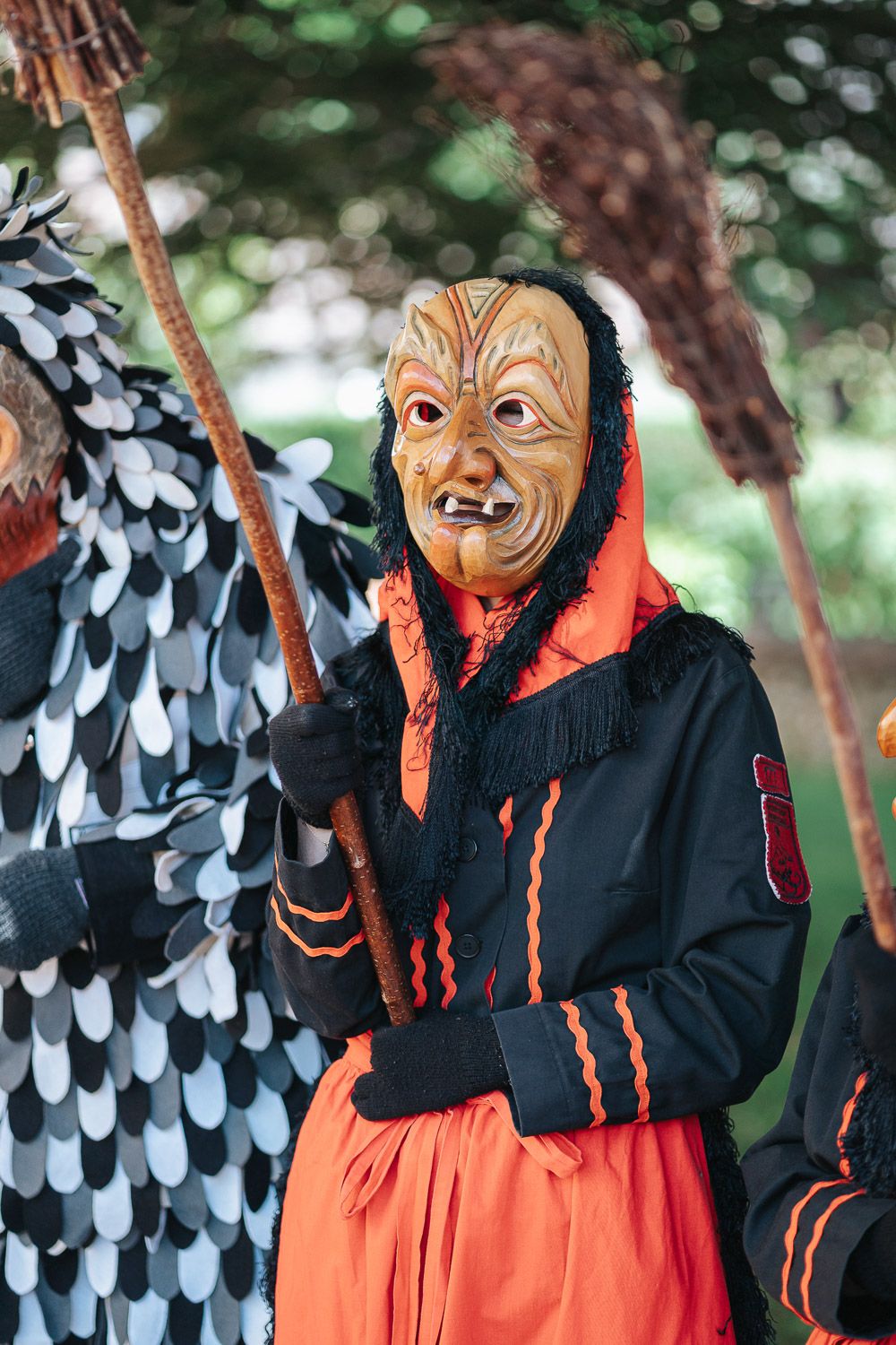 A person wearing an ornate mask and orange and black traditional costume stands among feathered displays outdoors.