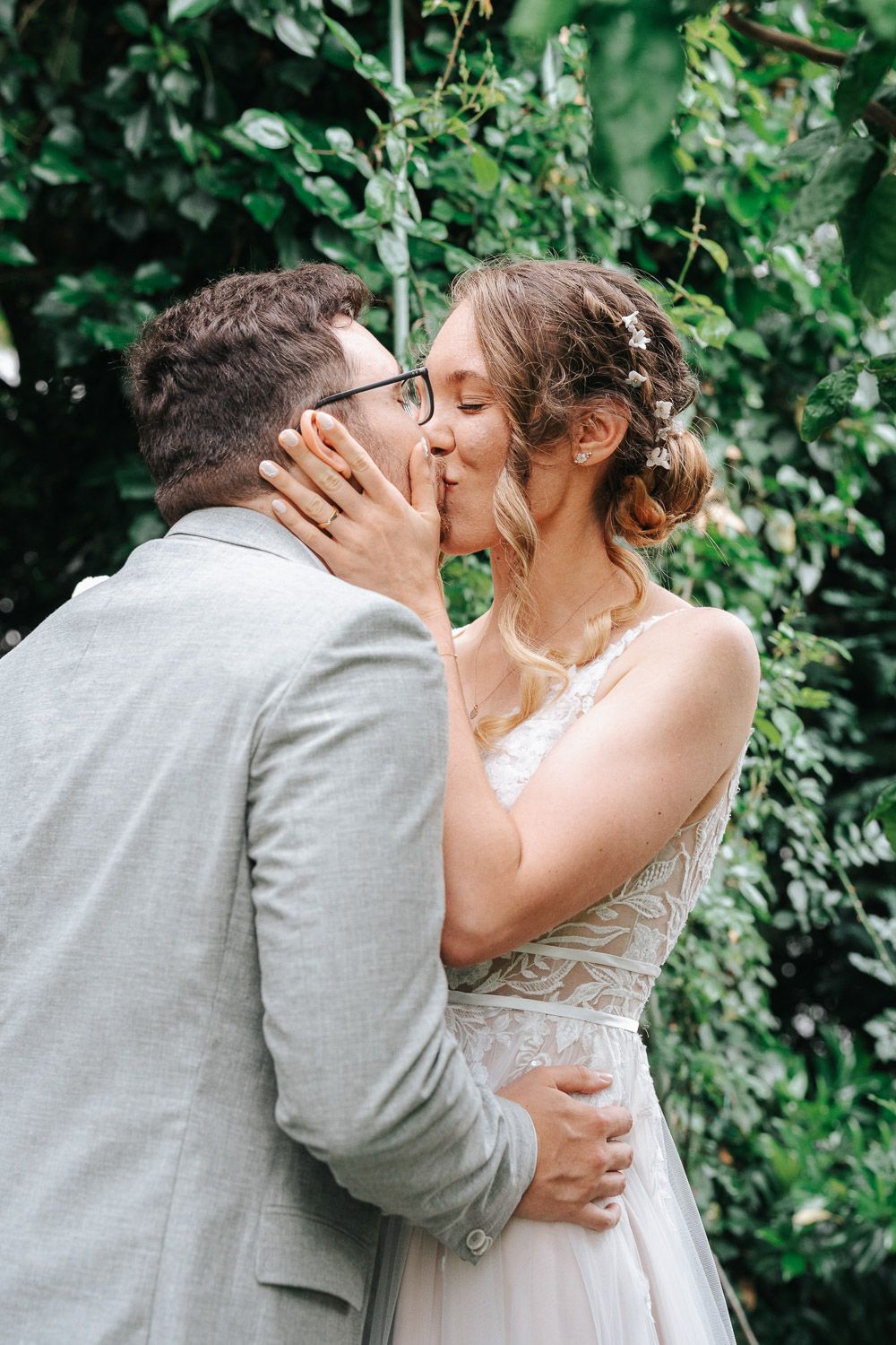 A romantic kiss shared between newlyweds in a garden setting with lush green foliage in the background.