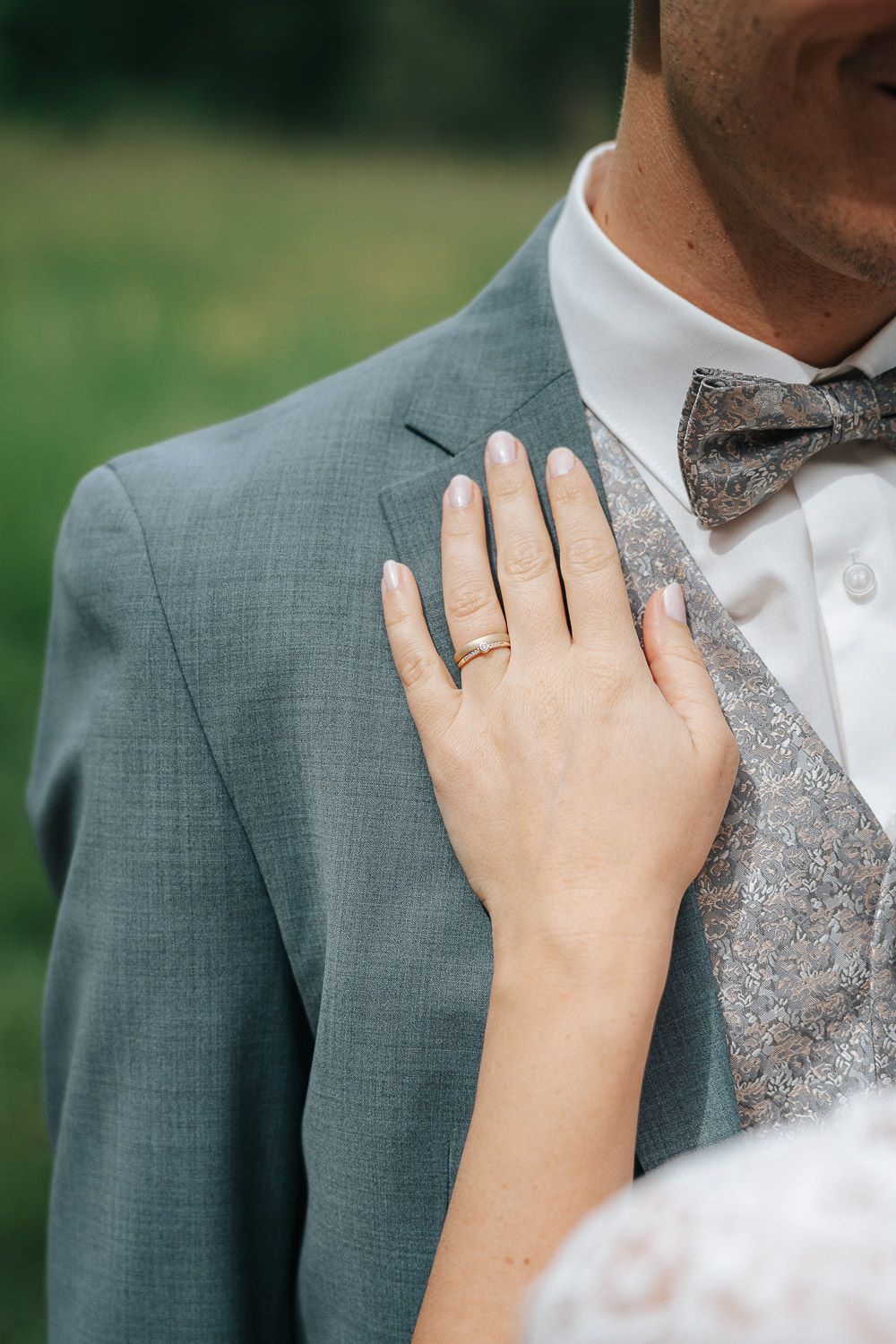 A delicate hand wearing a wedding ring rests on a gray suit jacket next to a patterned bowtie.