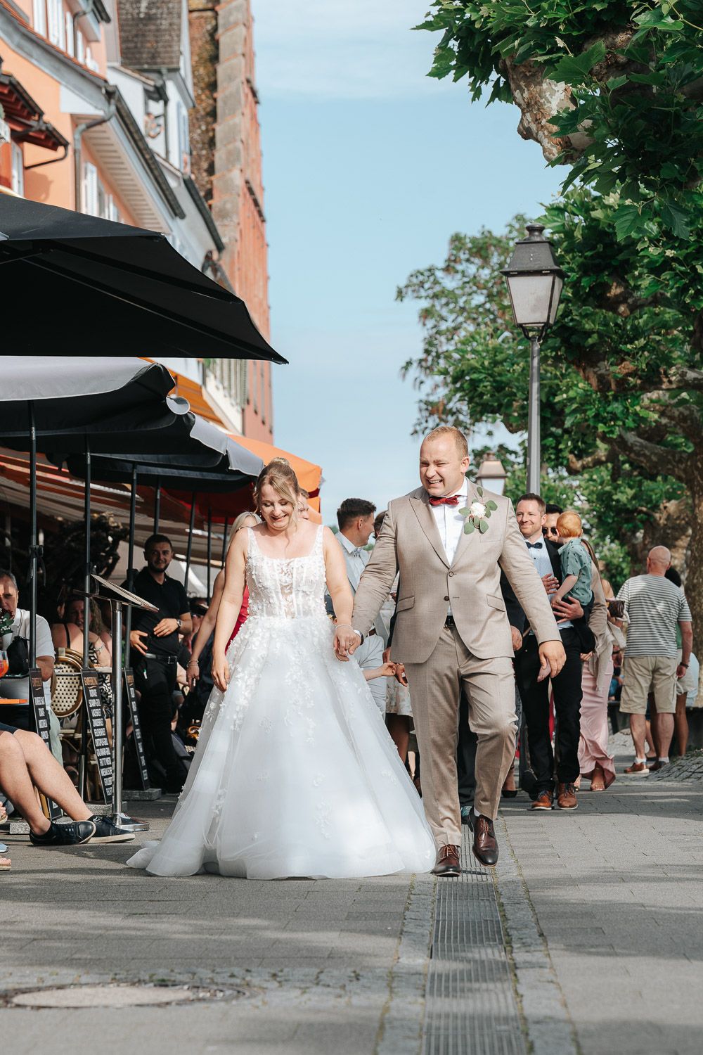 A wedding couple walking along a busy sidewalk outside shops with guests in the background during a sunny day.