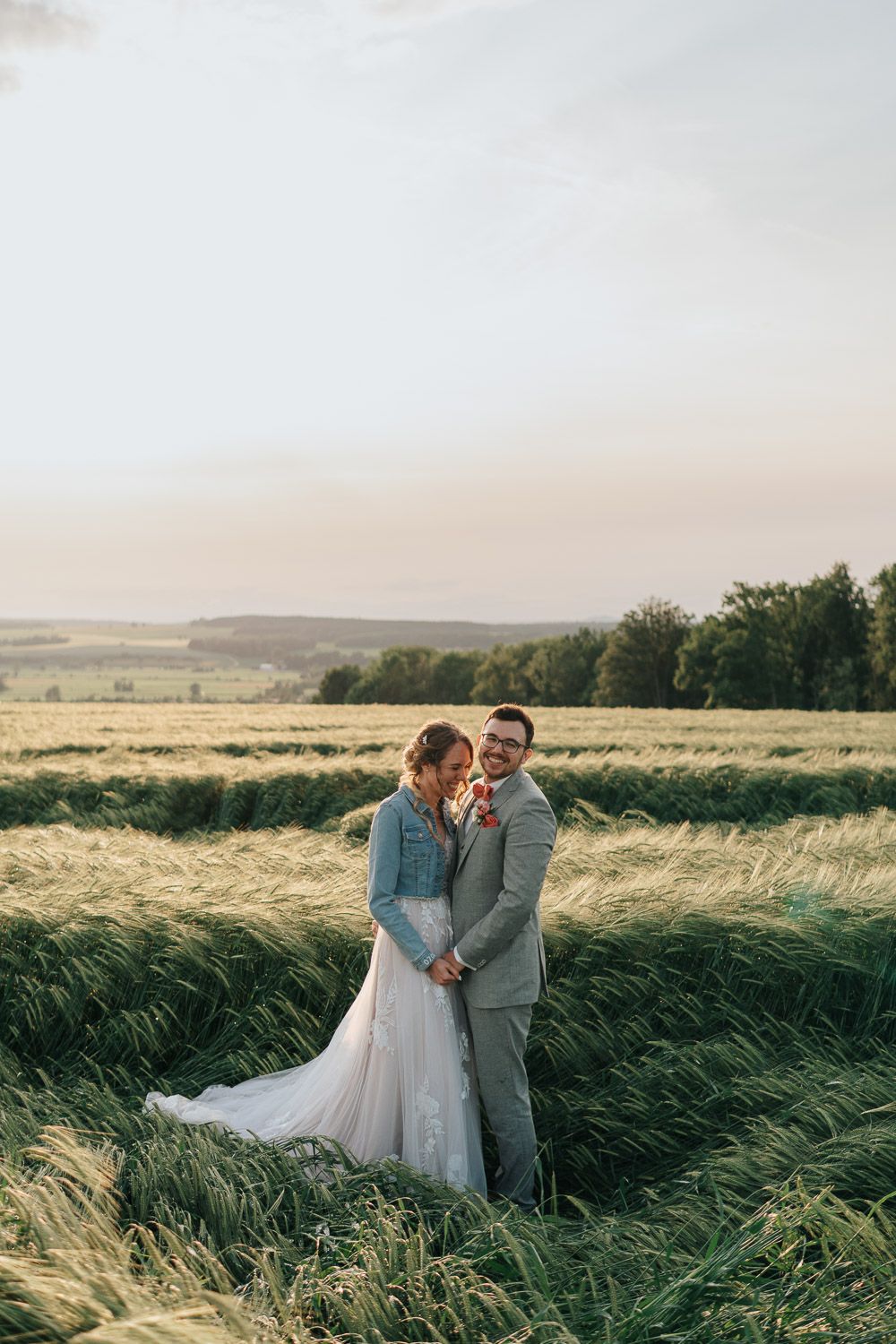 A couple stands together in flowing wheat field at sunset wearing wedding attire as wind blows through the tall grass.