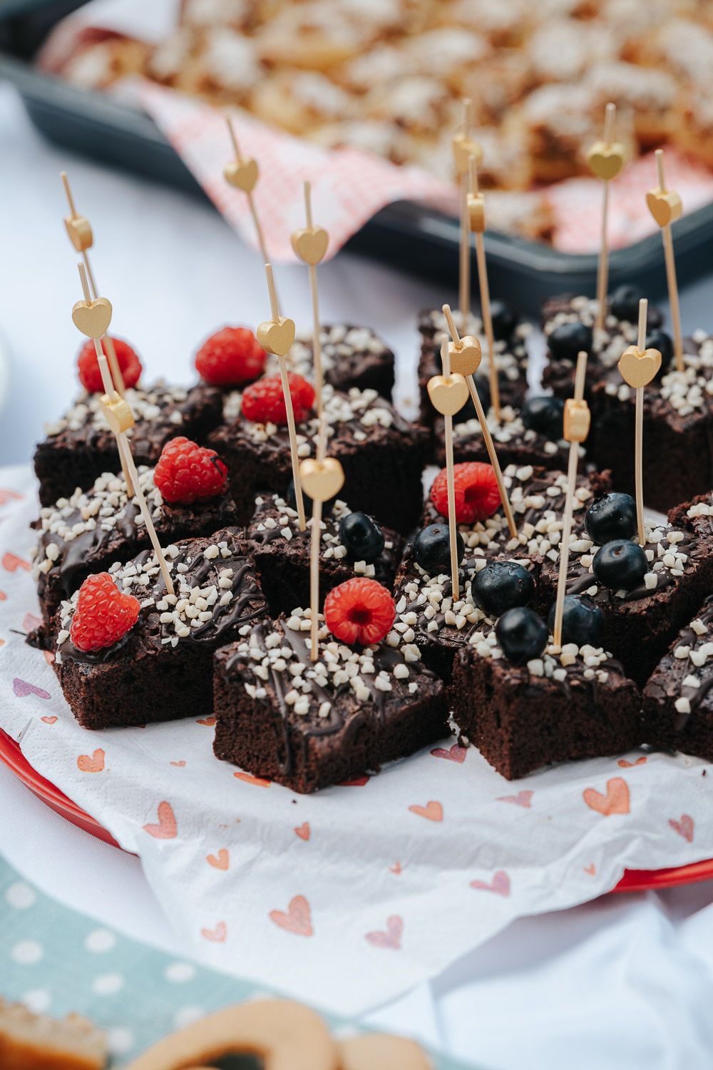 Chocolate brownies topped with chocolate sprinkles and fresh raspberries served on heart-patterned paper with decorative picks.