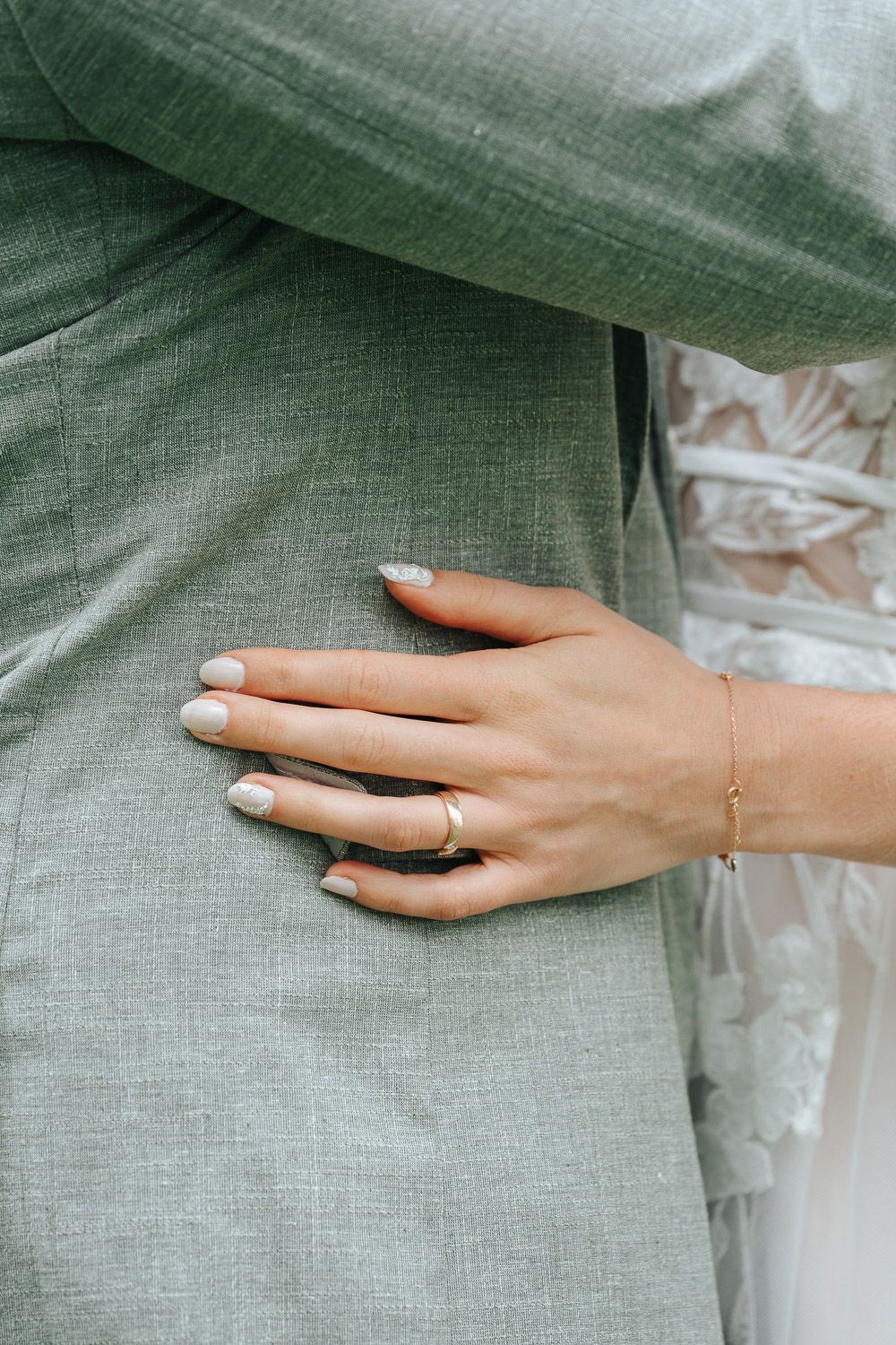 Close-up of a manicured hand with white nails and delicate bracelet against sage green fabric.