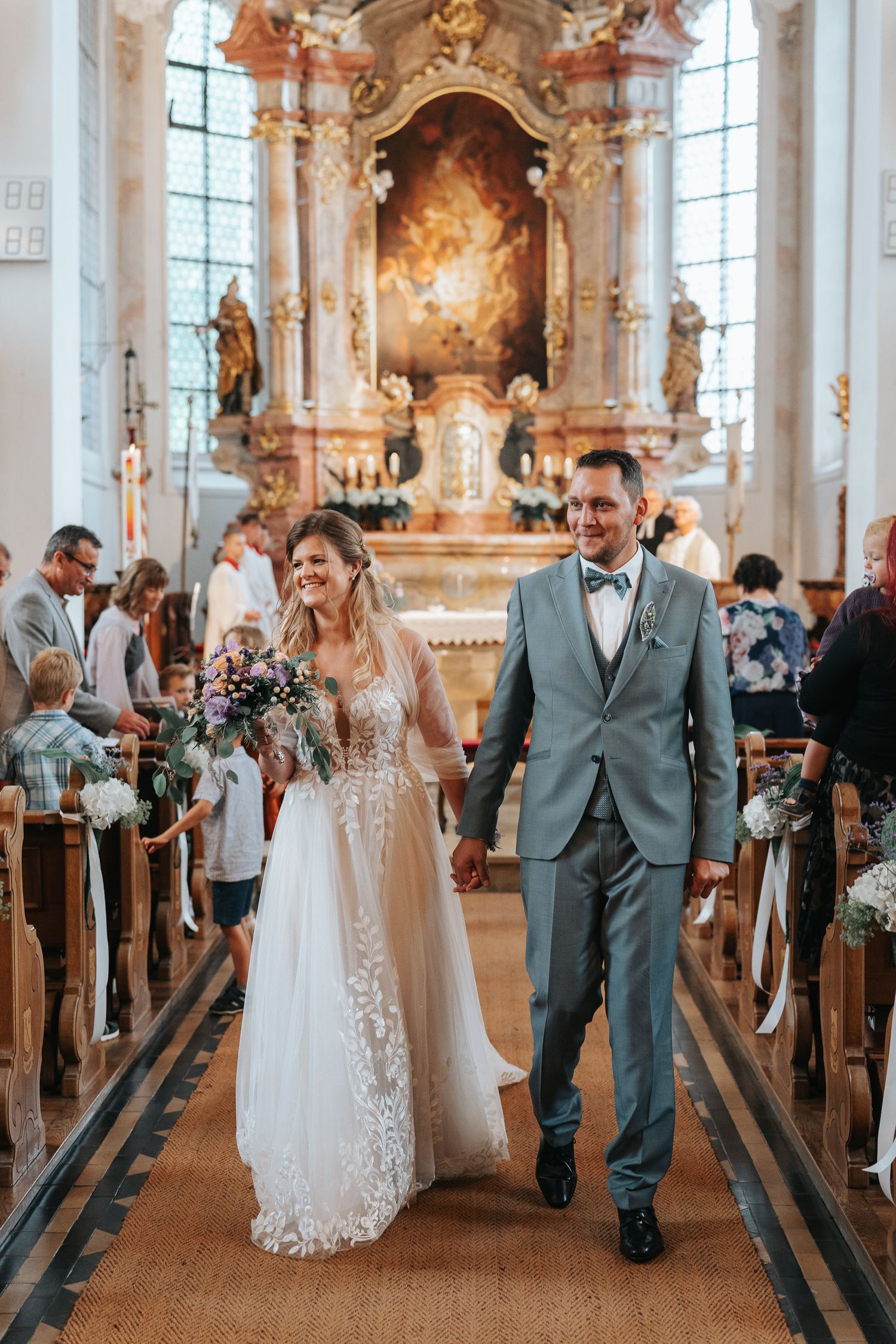 A wedding ceremony in an ornate baroque church with a decorated altar and wooden pews lining the aisle.