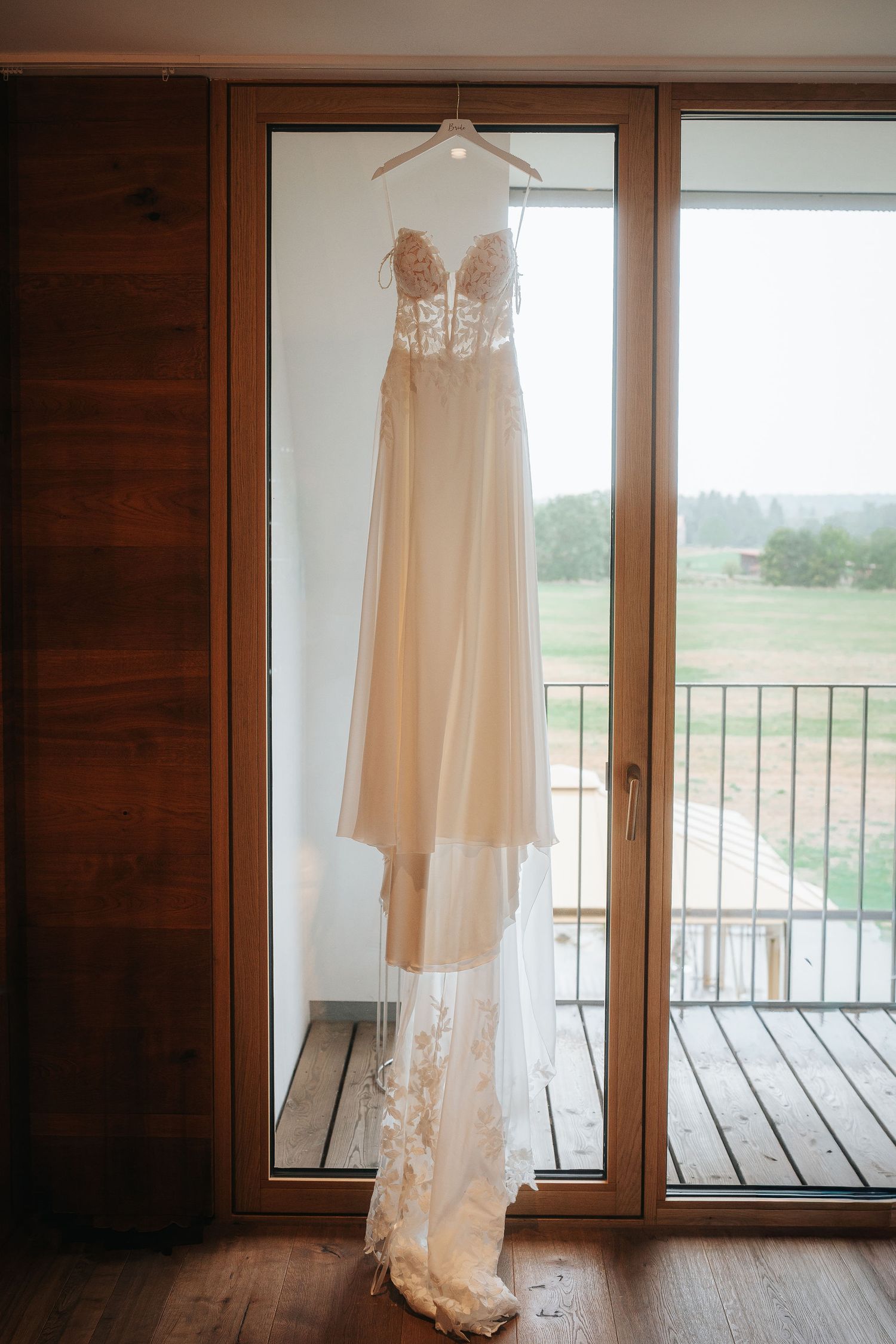 Elegant ivory lace wedding gown with a long flowing train hangs on a hanger in front of a glass balcony door overlooking green fields.