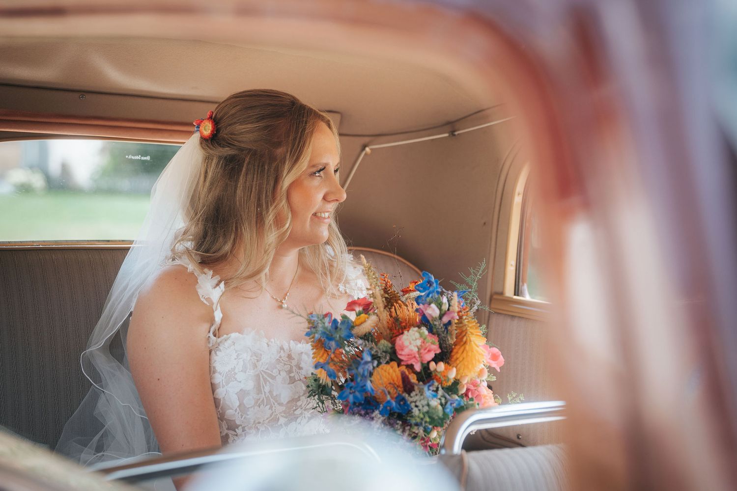Smiling bride in a lace wedding dress and veil sits in a vintage car holding a colorful bouquet of blue, orange and pink flowers.