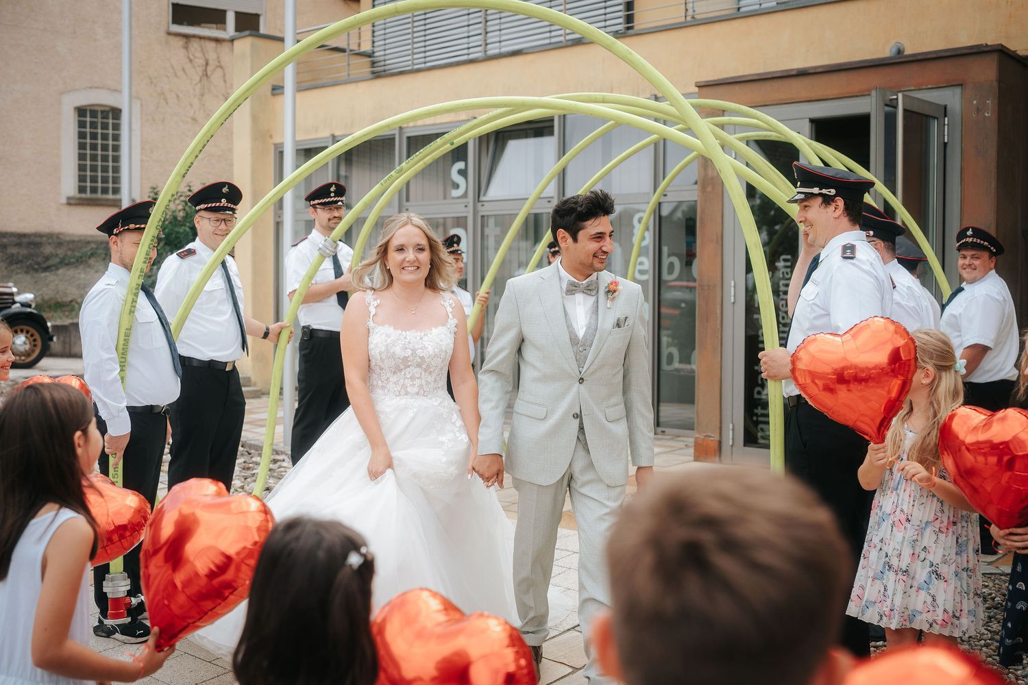 Bride and groom holding hands walk through a green arch past uniformed officers and children holding red heart-shaped balloons.